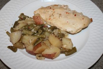 A white plate features a serving of baked chicken breast reminiscent of a Crock Pot Italian Chicken Recipe, alongside roasted green beans and chunks of red and white potatoes. The chicken is lightly seasoned with visible herbs, and the plate rests on a light-colored countertop.