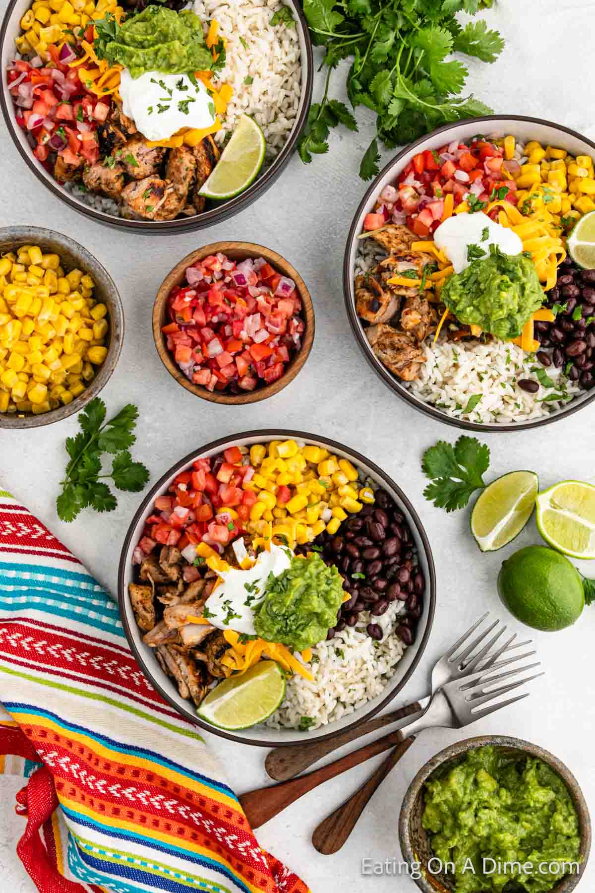 Grilled chicken in a bowl with diced tomatoes, corn, black beans and rice topped with guacamole and sour cream with shredded cheese with multiple bowls of this ingredients