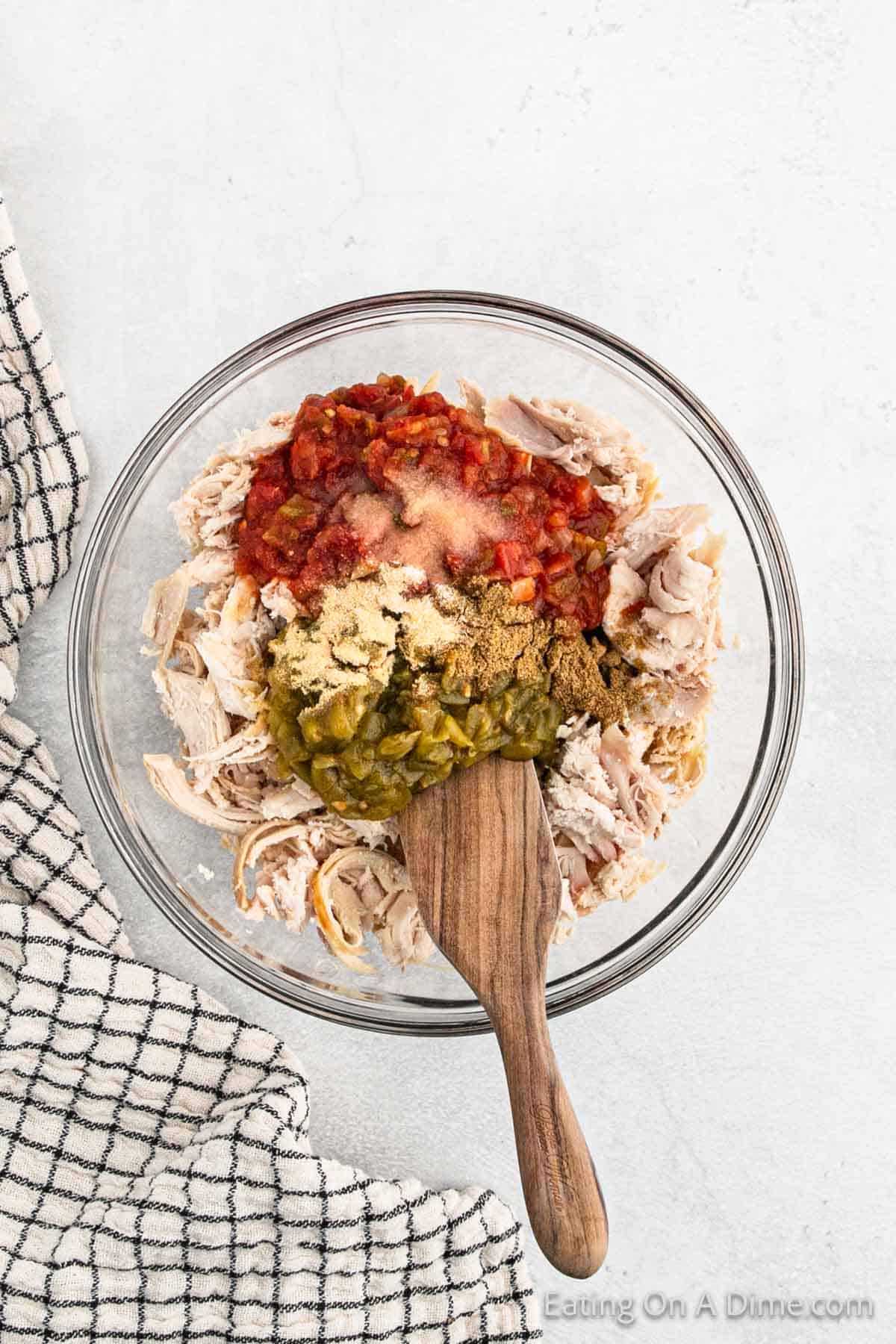 A glass mixing bowl contains shredded chicken topped with various ingredients such as diced tomatoes, green chilies, and spices, perfect for making a delicious Mexican Chicken Sandwich. A wooden spoon rests in the bowl. A black and white checkered cloth is partially visible on the left side of the image.