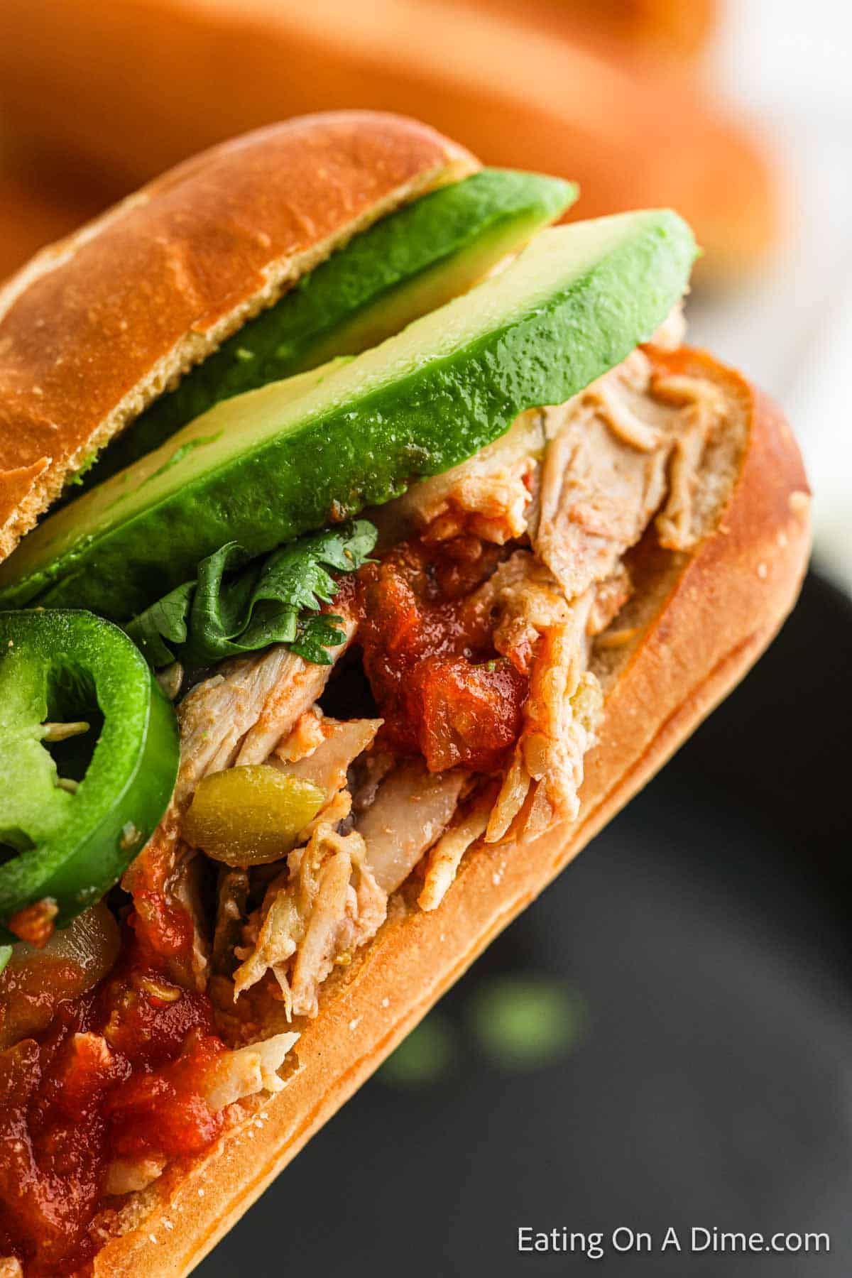 Close-up of a Mexican Chicken Sandwich filled with shredded chicken, salsa, spinach, avocado slices, and jalapeños in a hoagie roll. Text on the image reads "Eating On A Dime.com" in the bottom right corner.