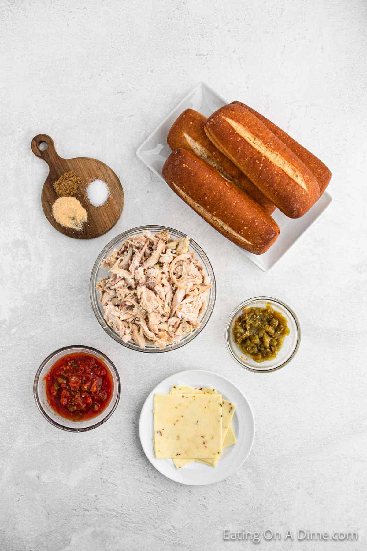 Ingredients for a Mexican Chicken Sandwich are laid out on a light gray surface. These include two hoagie rolls, a bowl of shredded chicken, a small bowl of salsa, a small bowl of relish, and a plate with slices of cheese. Text at the bottom reads "Eating On A Dime.com.