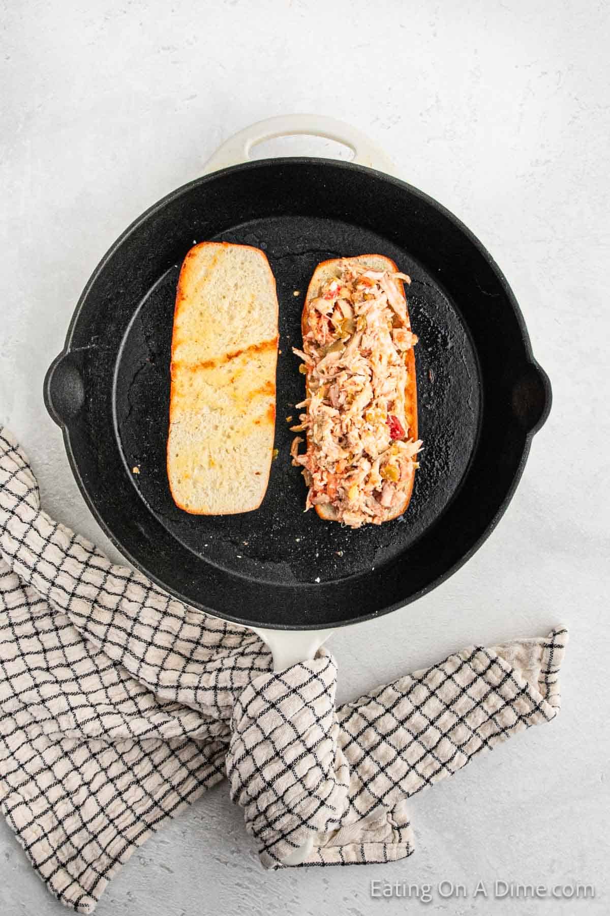 A cast iron skillet with an open Mexican Chicken Sandwich inside. One half of the sandwich is topped with a zesty shredded chicken mixture, while the other half is plain. A checkered towel is placed beside the skillet on a light-colored surface.