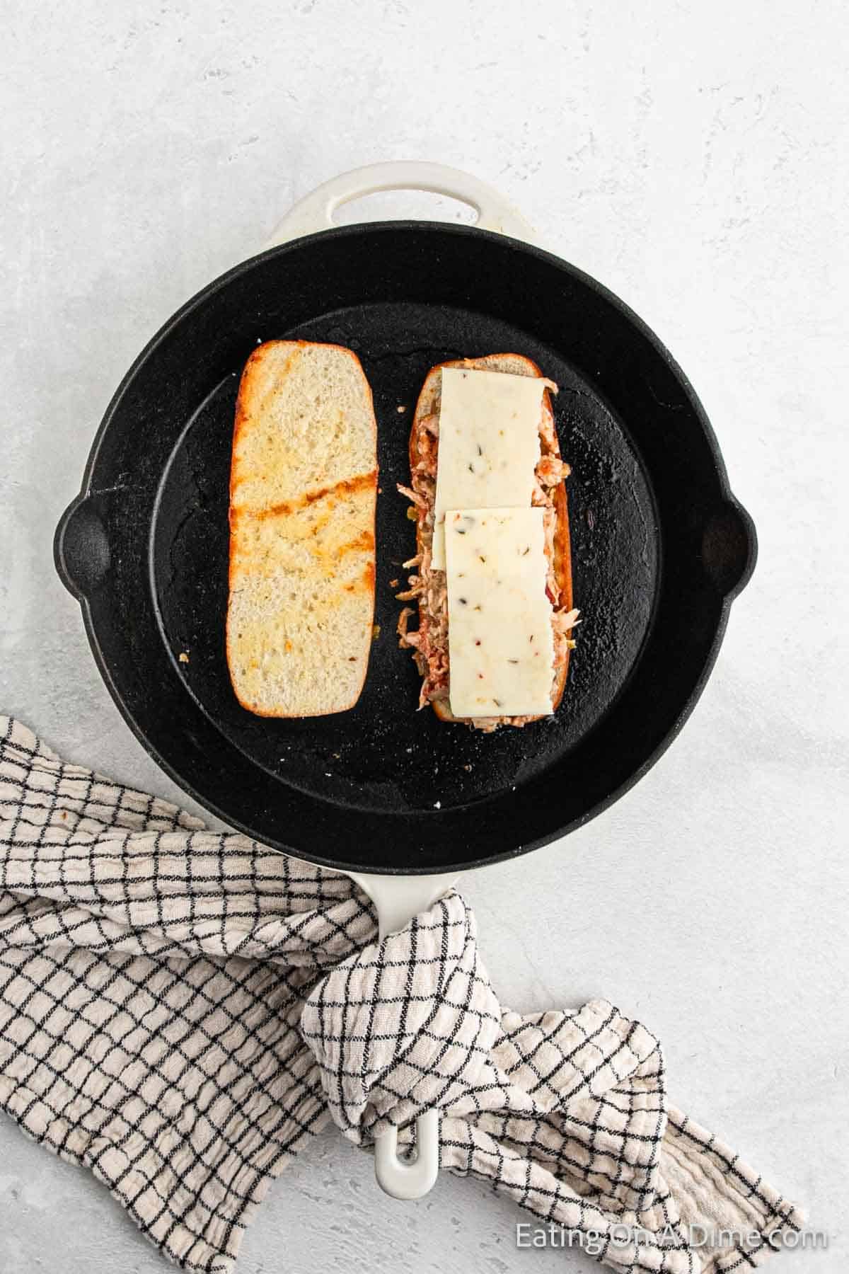 A Mexican Chicken Sandwich is being prepared in a cast iron skillet. The sandwich has two halves; the left half is a toasted plain bun, while the right half boasts a savory chicken filling topped with two slices of white cheese. A black and white checkered cloth is wrapped around the skillet handle.