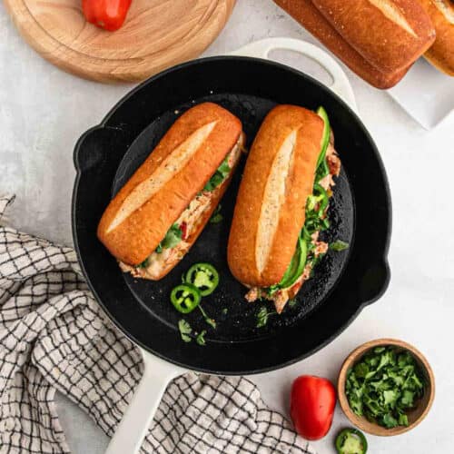 A black skillet contains two stuffed baguettes resembling a delightful Mexican Chicken Sandwich, with visible meat, vegetables, and fresh greens. Nearby, a wooden surface holds a sliced red bell pepper, a bowl of chopped cilantro, a checkered cloth, more baguettes, and another red bell pepper.