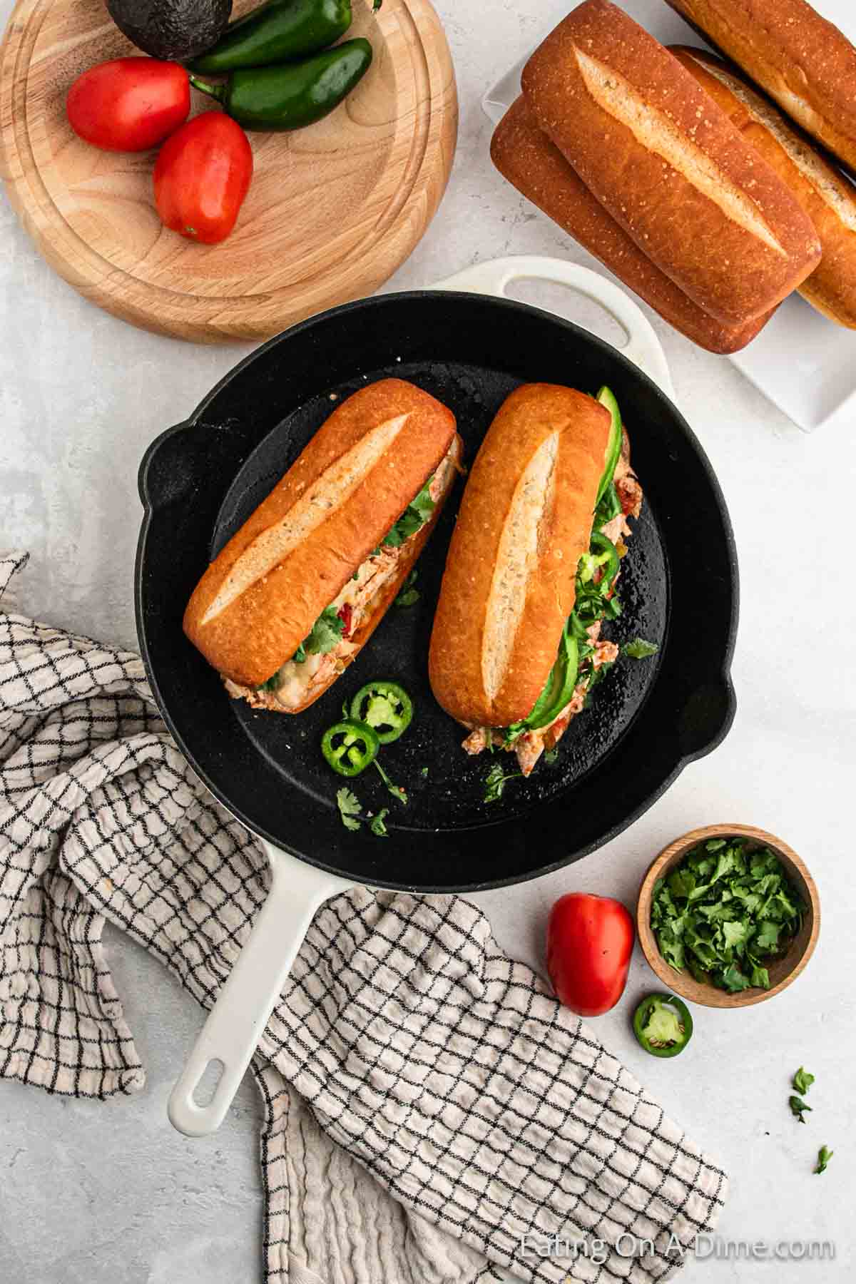 A white skillet on a countertop holds two Mexican Chicken Sandwiches with crusty bread, filled with vegetables and what appears to be meat. Surrounding the skillet are halved tomatoes, jalapeños, a bowl of chopped herbs, a wooden cutting board with sandwich rolls, and a kitchen towel.