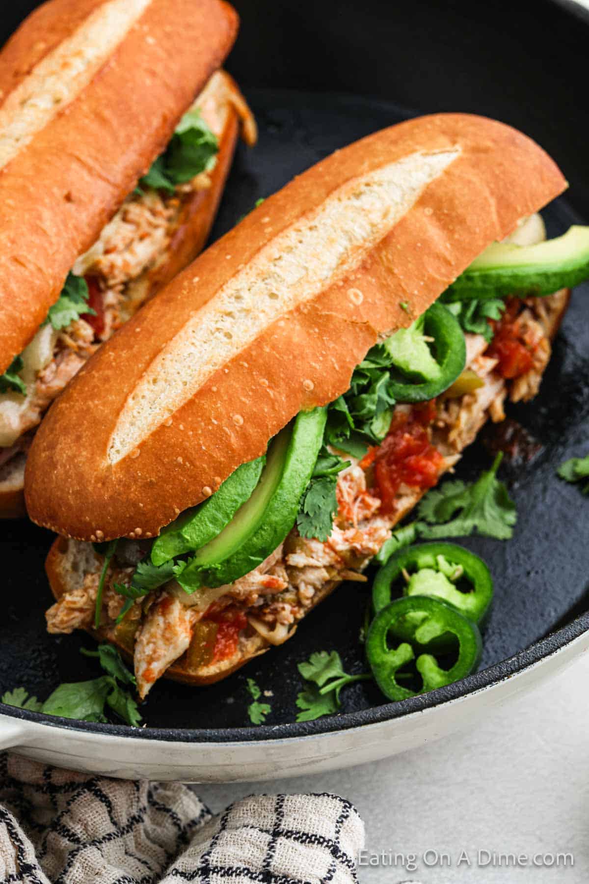 Two Mexican chicken sandwiches filled with shredded chicken, fresh cilantro, jalapeno slices, and other ingredients are placed in a black skillet. The bread is toasted with a scored top. A checkered napkin is partially visible at the bottom of the skillet.