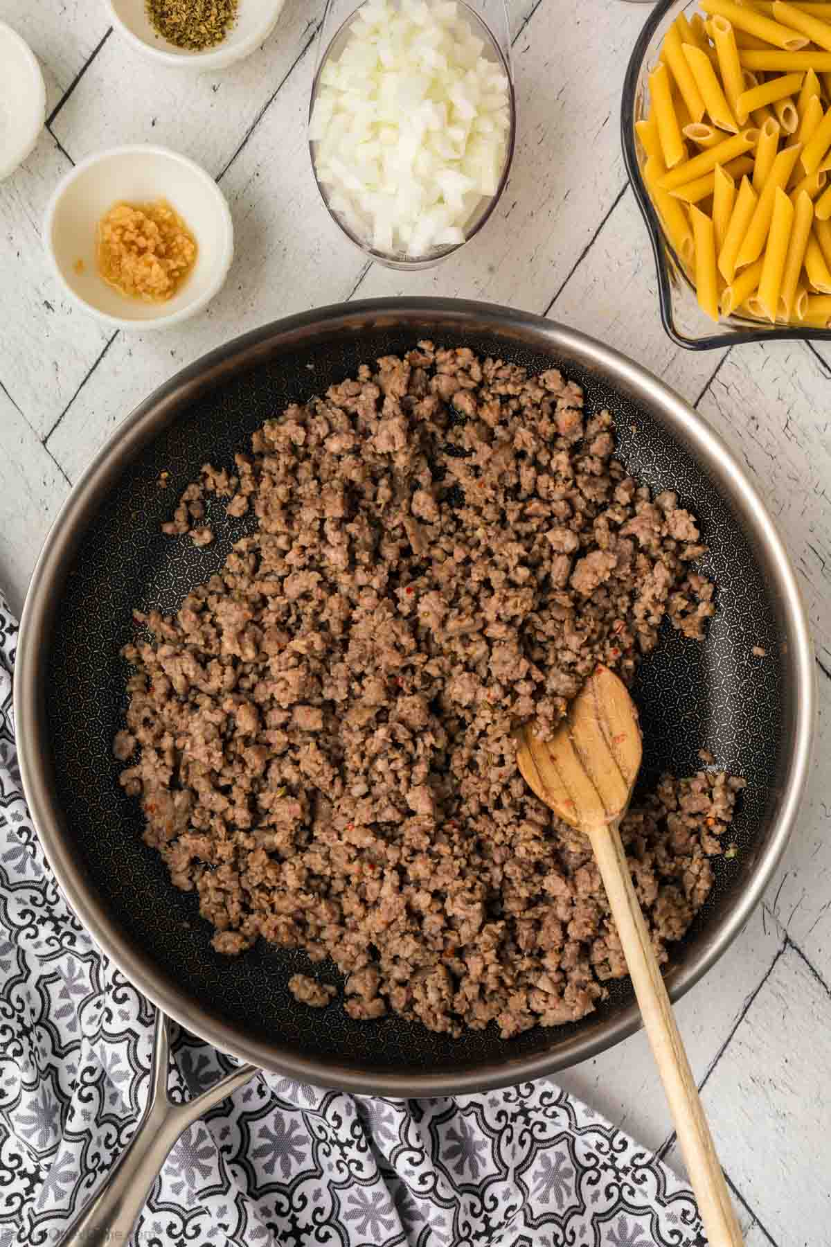 Cooked ground beef in a skillet with a wooden spoon with a bowl of fresh onions and pasta with minced garlic and pepper