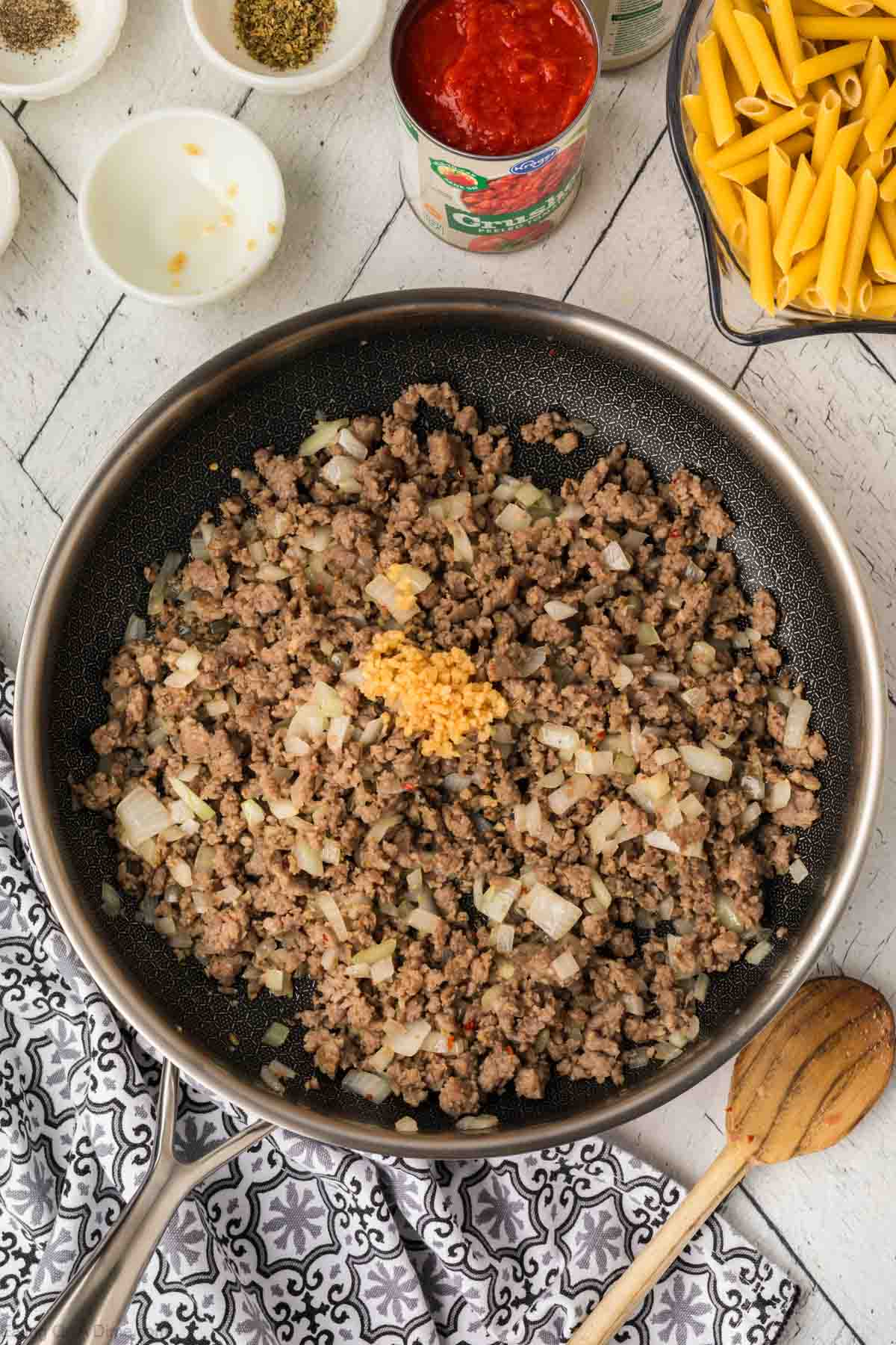 Adding minced garlic to skillet with ground beef with fresh onions and a side of pasta and canned tomatoes