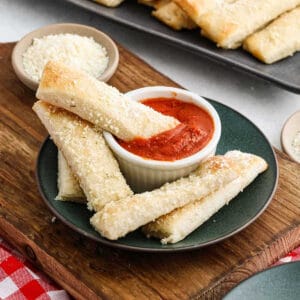 A plate of Little Caesars Crazy Bread sprinkled with grated cheese surrounds a small dish of marinara sauce on a rustic wooden board. Additional cheese graces the backdrop, complementing the red and white checkered cloth setting.