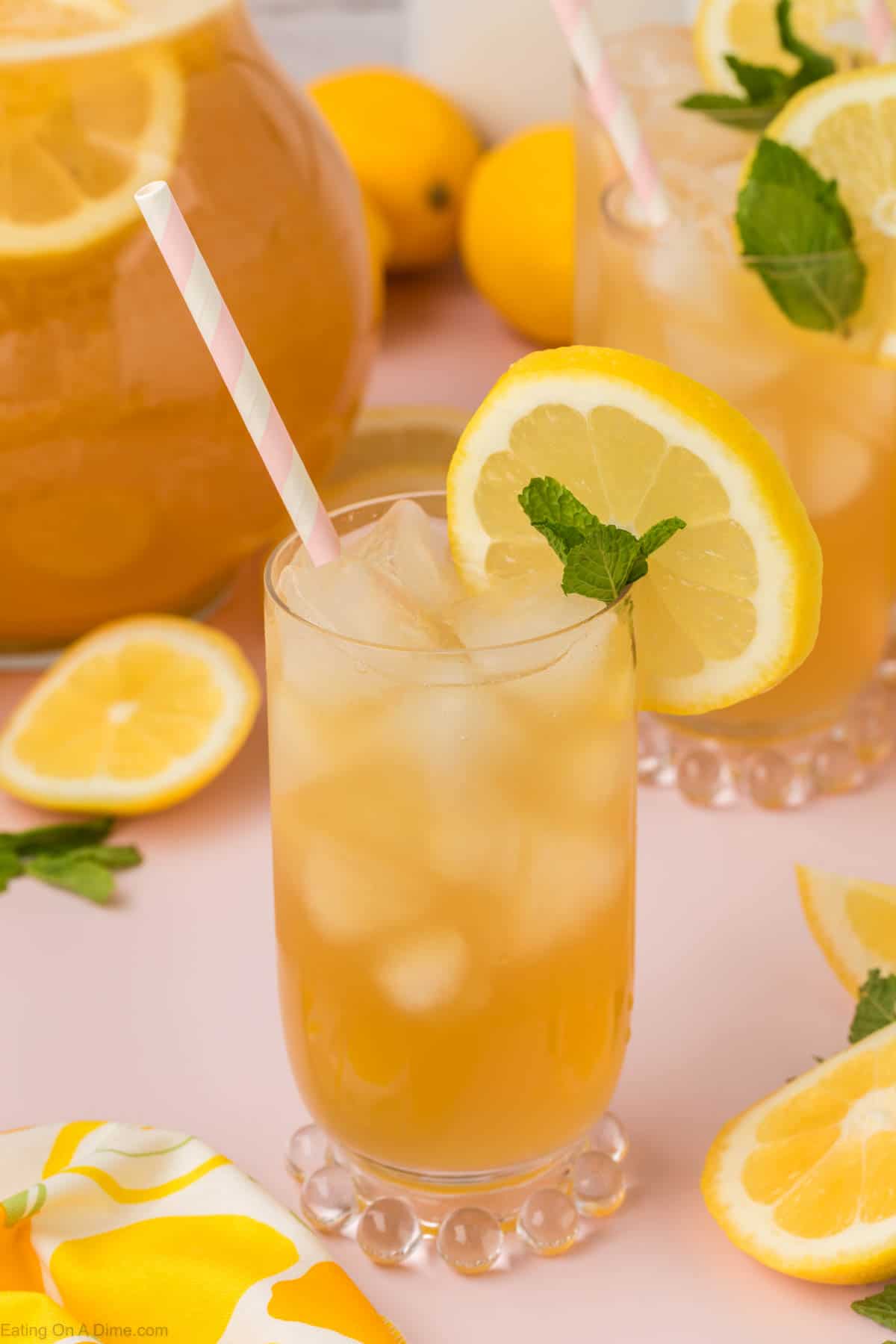 Glass of sparkling lemonade in a glass garnished with a lemon slice and fresh mint. Pitcher of lemonade in the background