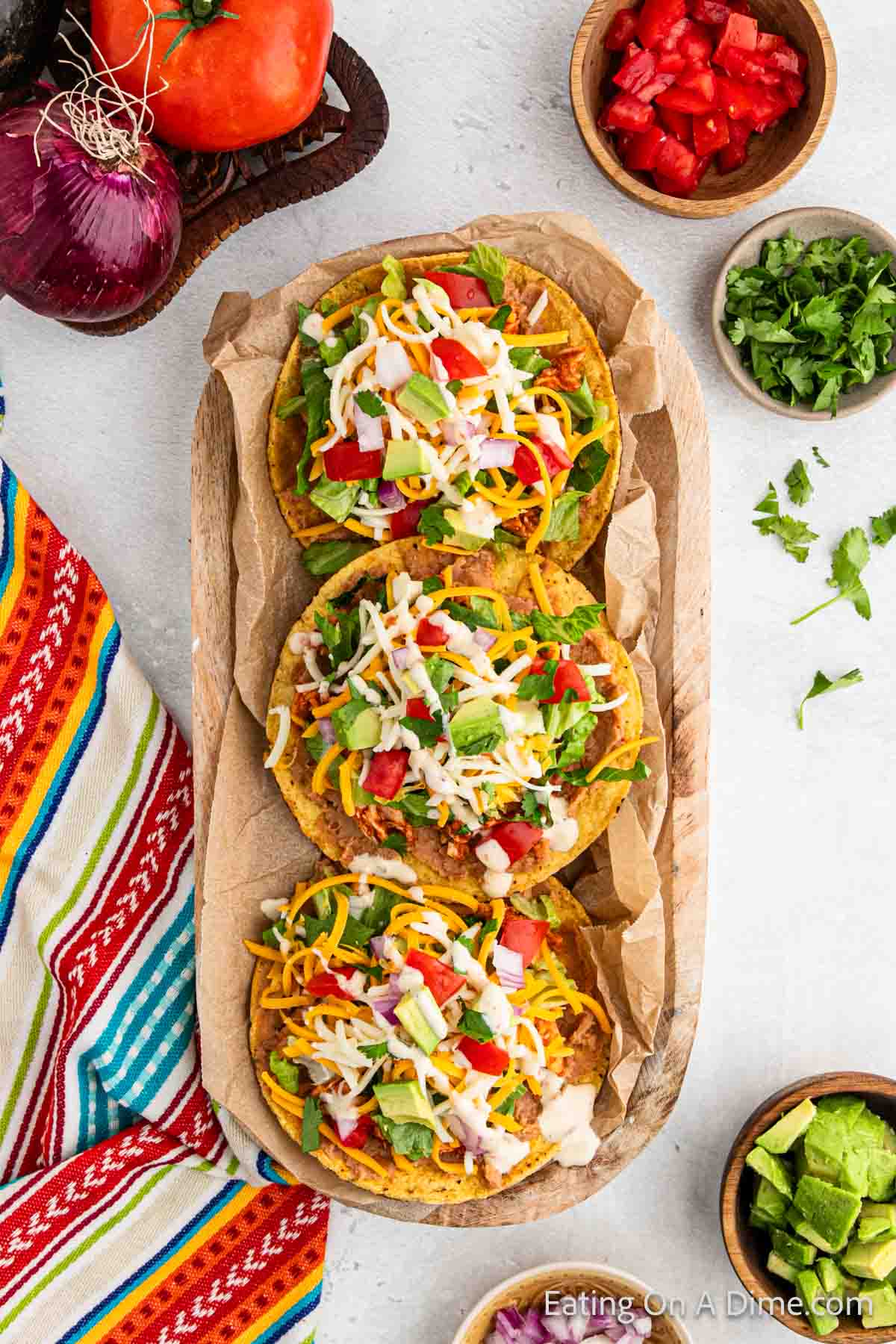 Chicken Tostadas on a platter with a platter with fresh red onions and tomatoes, a bowl of diced tomatoes, cilantro, diced avocado and red onions on the side