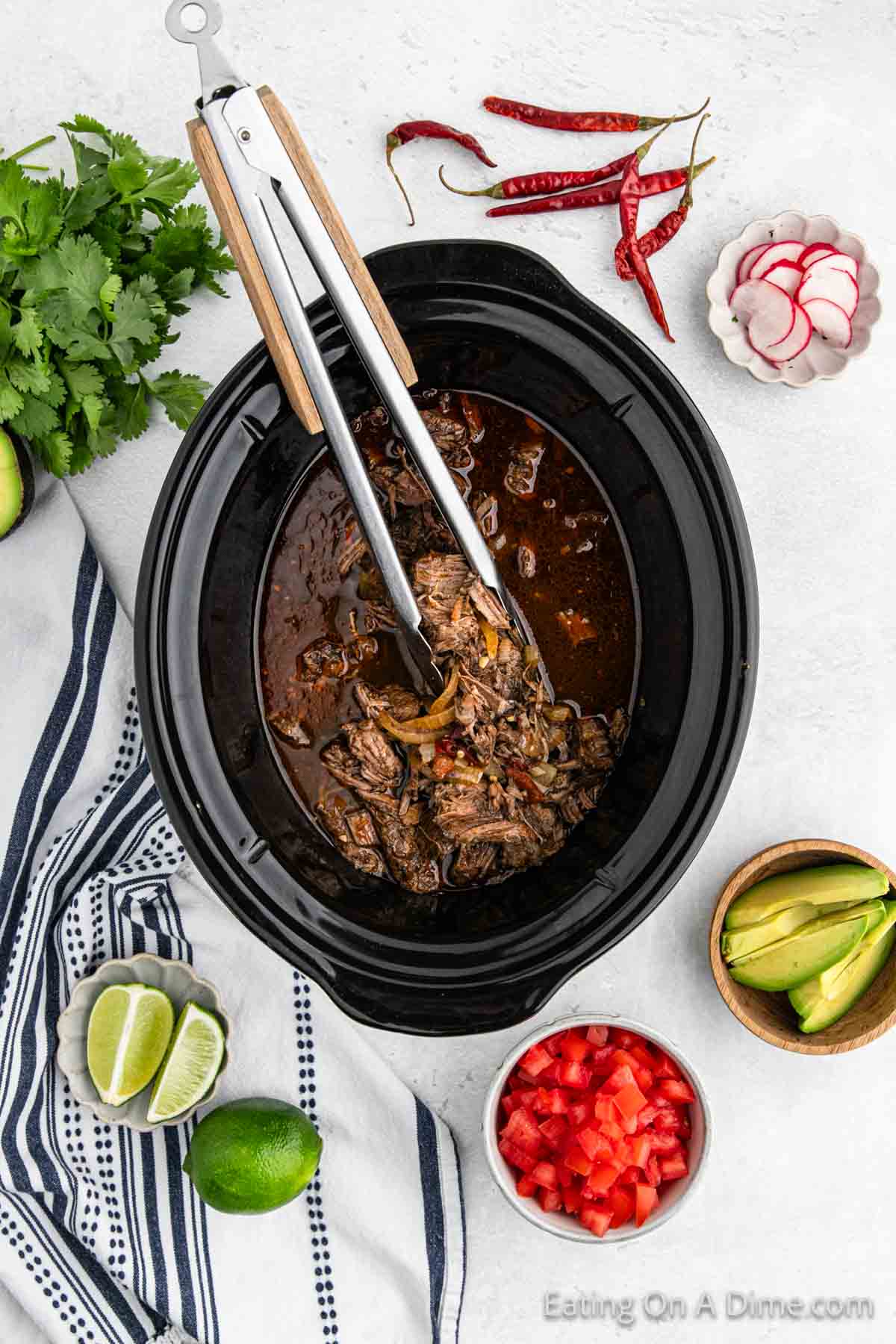 Shredded meat in slow cooker with silver tongs with bowls of avocado, diced tomato, and lime wedges