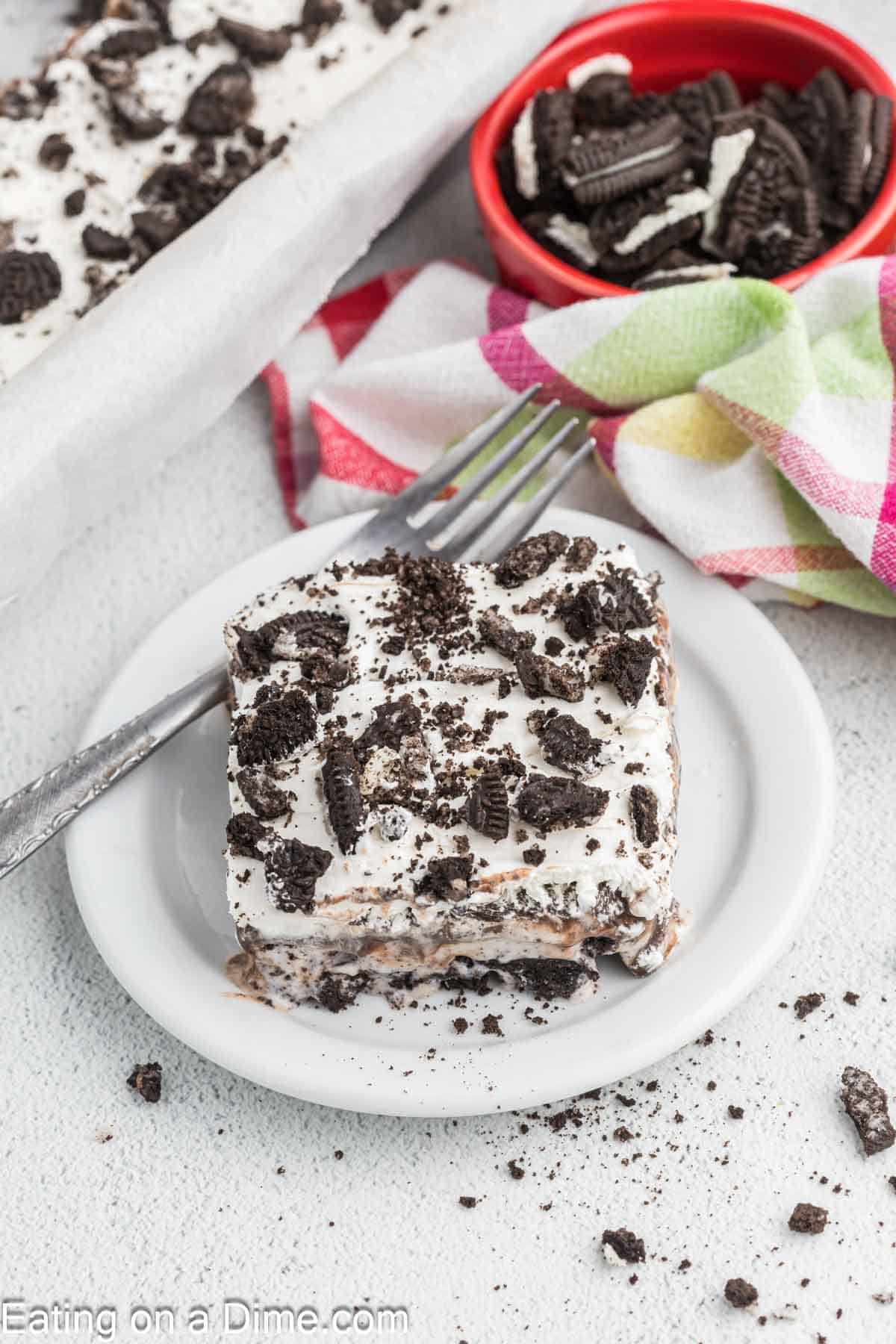 Slice of Cookies and Cream Ice Cream Cake on a plate with a fork with a small bowl of crushed Oreos