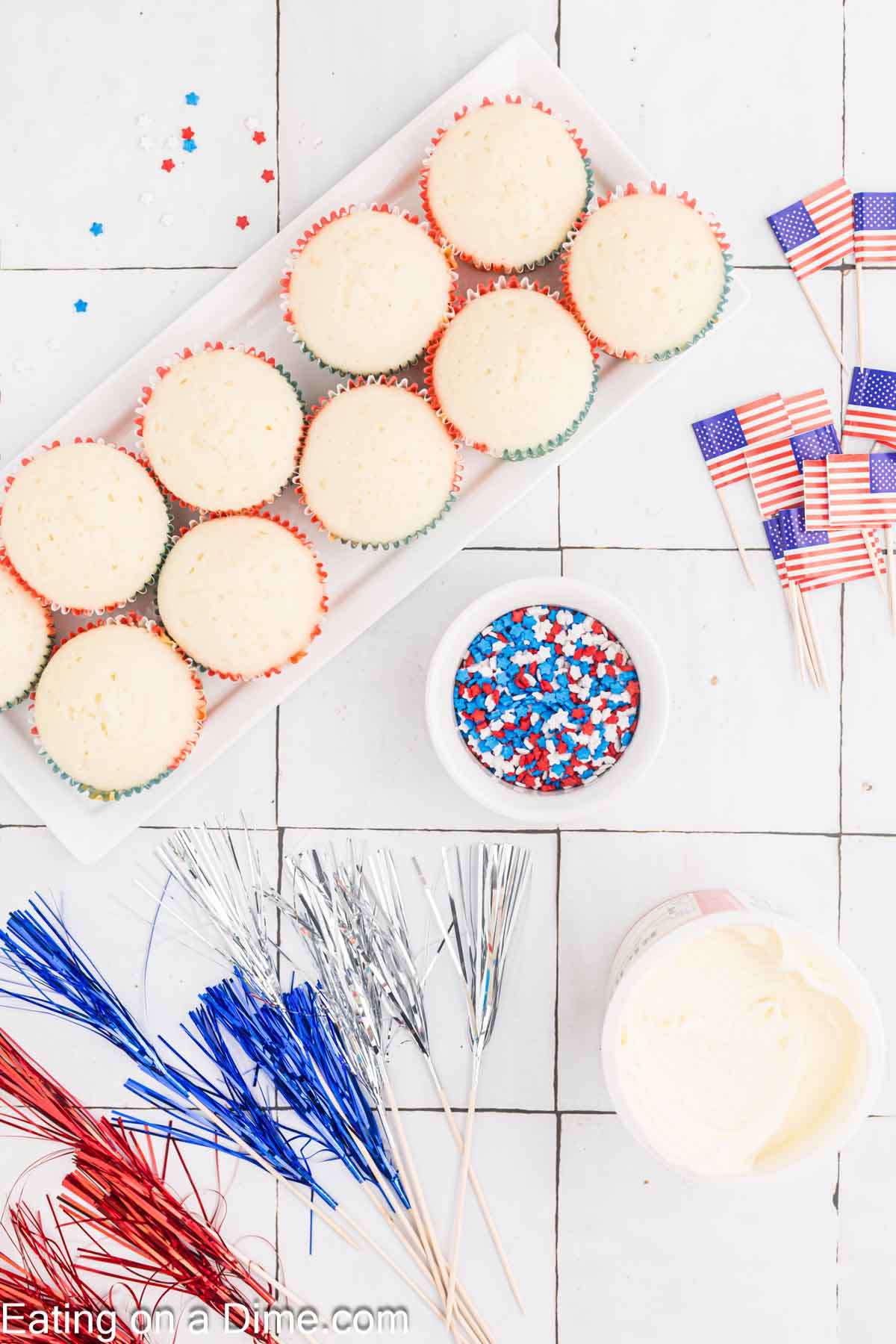A tray of unfrosted Firecracker Cupcakes sits on a tile countertop, surrounded by small American flag toothpick toppers, red, white, and blue star-shaped sprinkles in a bowl, a tub of white frosting, and metallic blue, red, and silver decorations.
