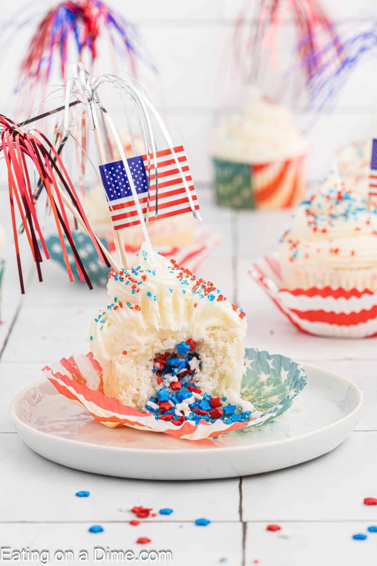 A festive Firecracker Cupcake with red, white, and blue sprinkles and an American flag pick sits on a plate. The cupcake wrapper is open, revealing a bite taken out of the cupcake, exposing more red, white, and blue sprinkles inside. Other decorated cupcakes are in the background.