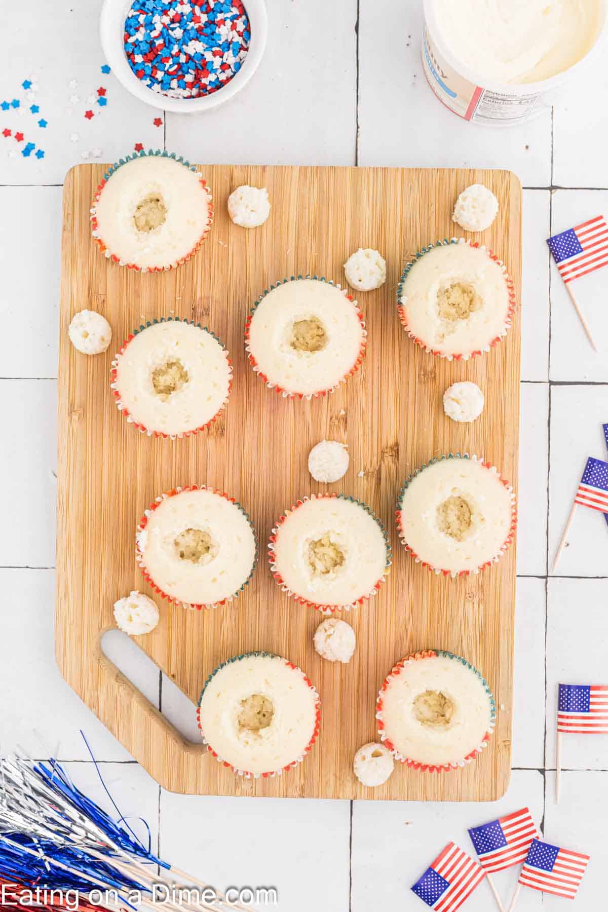 A wooden board displays nine Firecracker Cupcakes with holes cut in the center. A small knife rests on the board. Surrounding the board are a container of white frosting, a bowl of blue, white, and red sprinkles, mini American flags, and patriotic decorations.