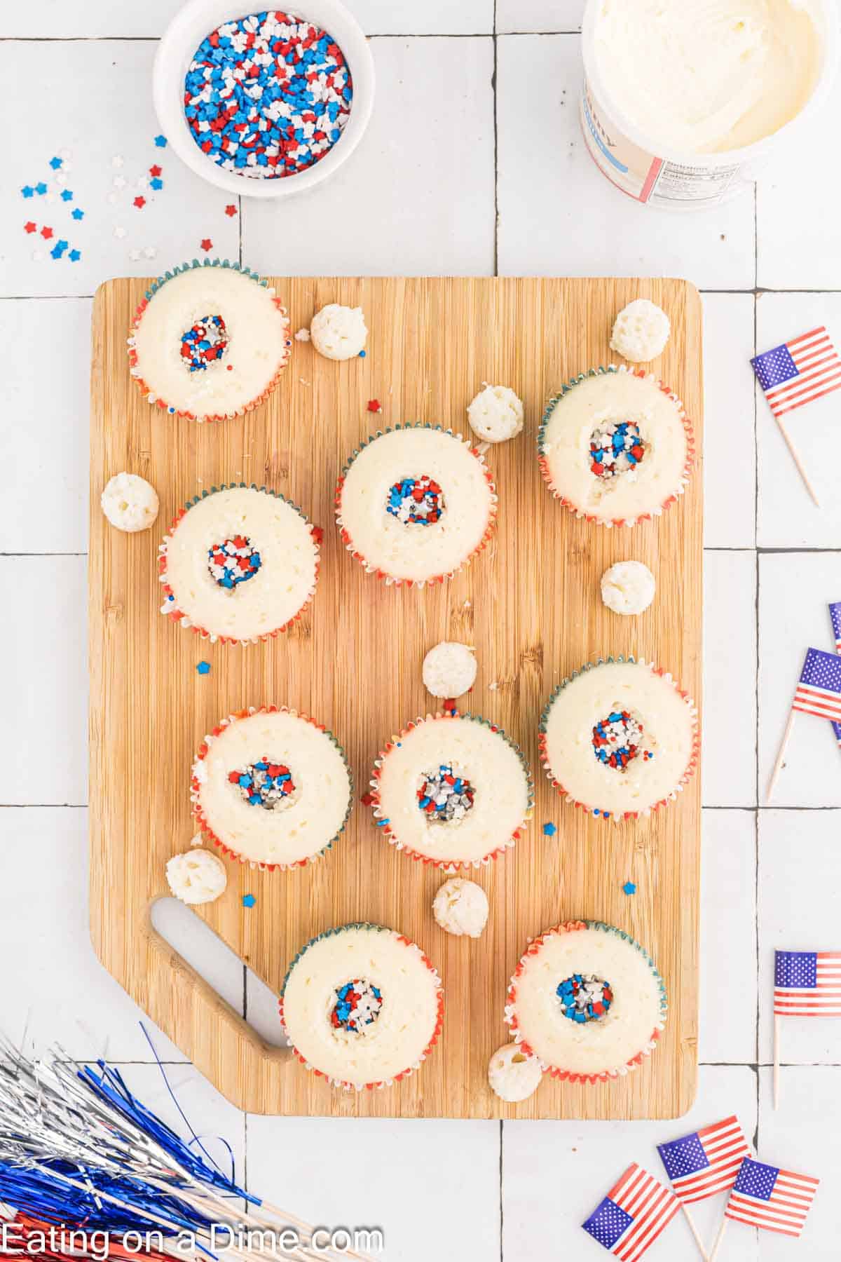 A wooden cutting board displays nine Firecracker Cupcakes frosted with white icing and topped with red, white, and blue star-shaped sprinkles. An icing spatula, a bowl of sprinkles, a tub of frosting, and small American flags adorn the white-tiled surface around the board.
