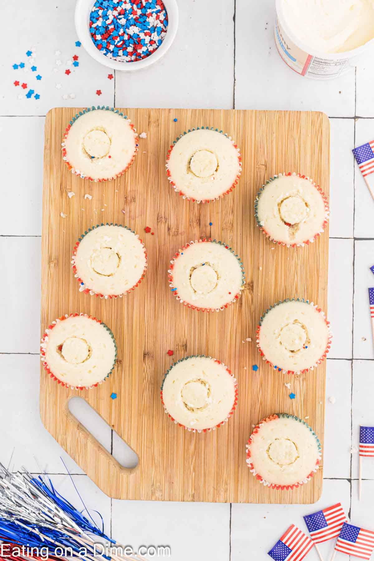 A wooden cutting board holds nine Firecracker Cupcakes with patriotic red, white, and blue sprinkles. A bowl of sprinkles, a container of frosting, and small American flag decorations are nearby, creating a festive atmosphere. The website “Eating on a Dime” is partially visible.