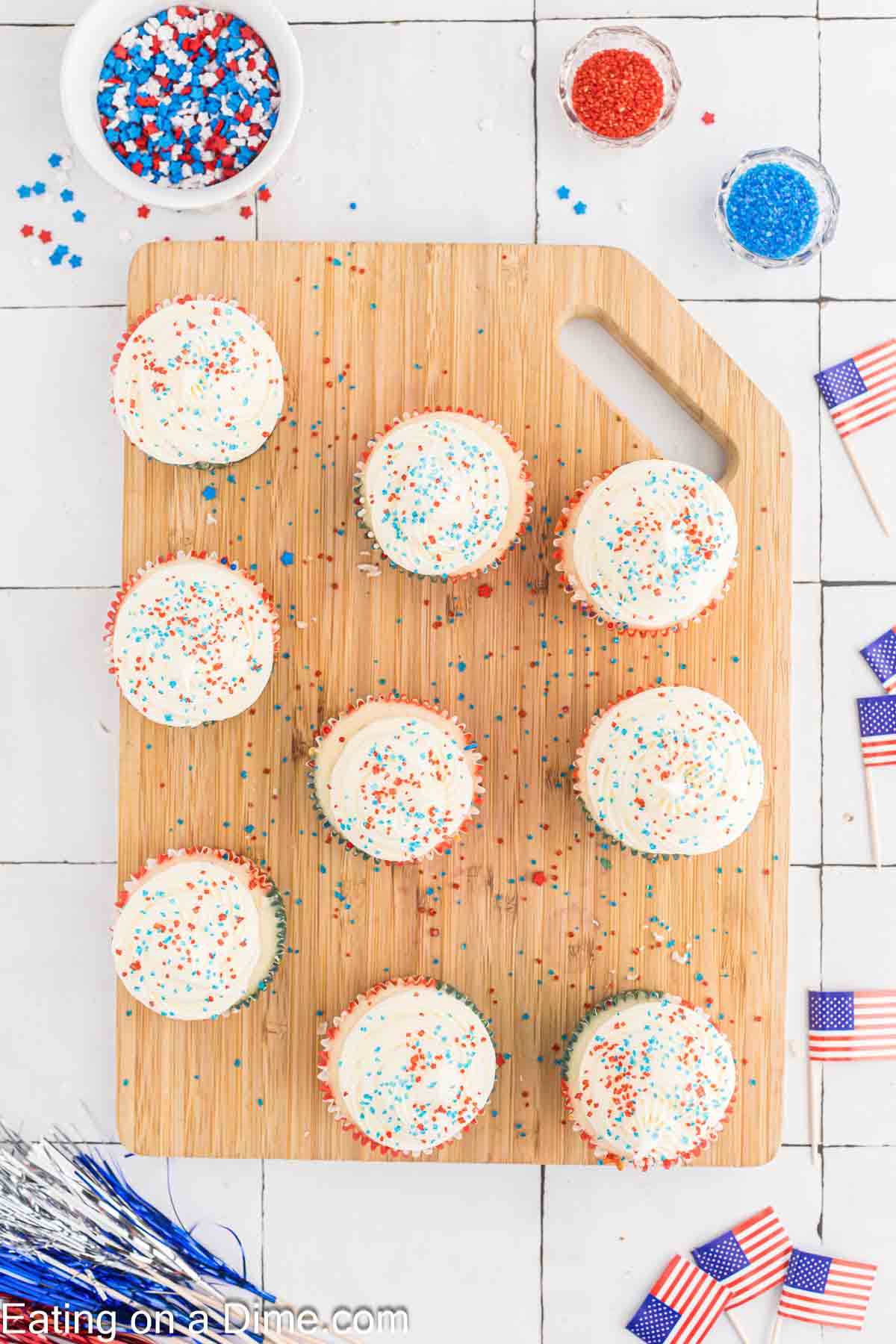 A wooden cutting board holds nine Firecracker Cupcakes topped with white frosting and red, white, and blue sprinkles. The table has bowls of sprinkles and American flags. The scene has a festive, patriotic theme.