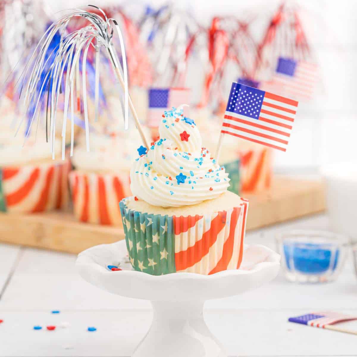 A festive Firecracker Cupcake topped with vanilla frosting, blue and red sprinkles, a small American flag, and a silver fringe decoration. The cupcake liner features a stars and stripes design. In the background, more similarly decorated cupcakes are visible.