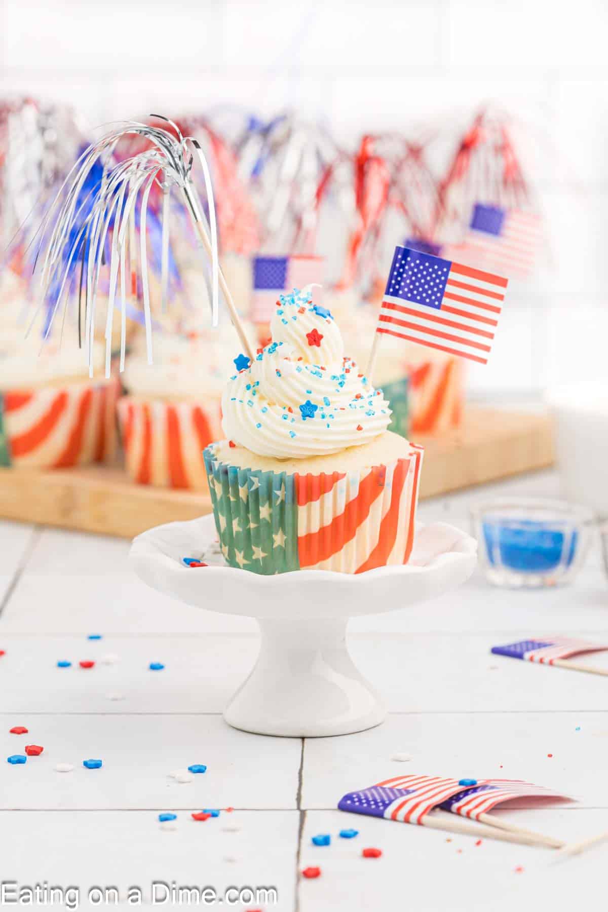 A festive Firecracker Cupcake with red, white, and blue swirl frosting, topped with star-shaped sprinkles, a small American flag, and silver tinsel sticks, displayed on a white stand. More similar cupcakes and patriotic decorations are visible in the background.