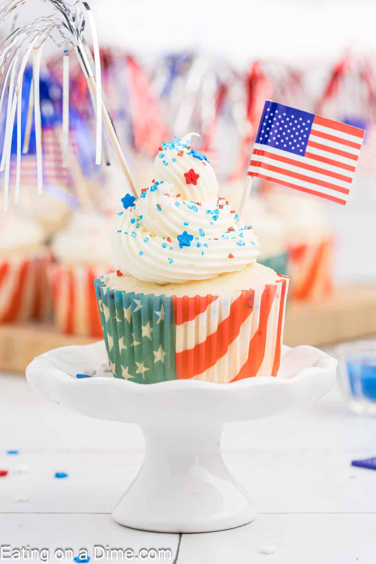 A patriotic Firecracker Cupcake with white frosting, blue and red star sprinkles, and a small American flag on top. The cupcake liner features the American flag design. It sits on a white cake stand with more decorated cupcakes blurred in the background.