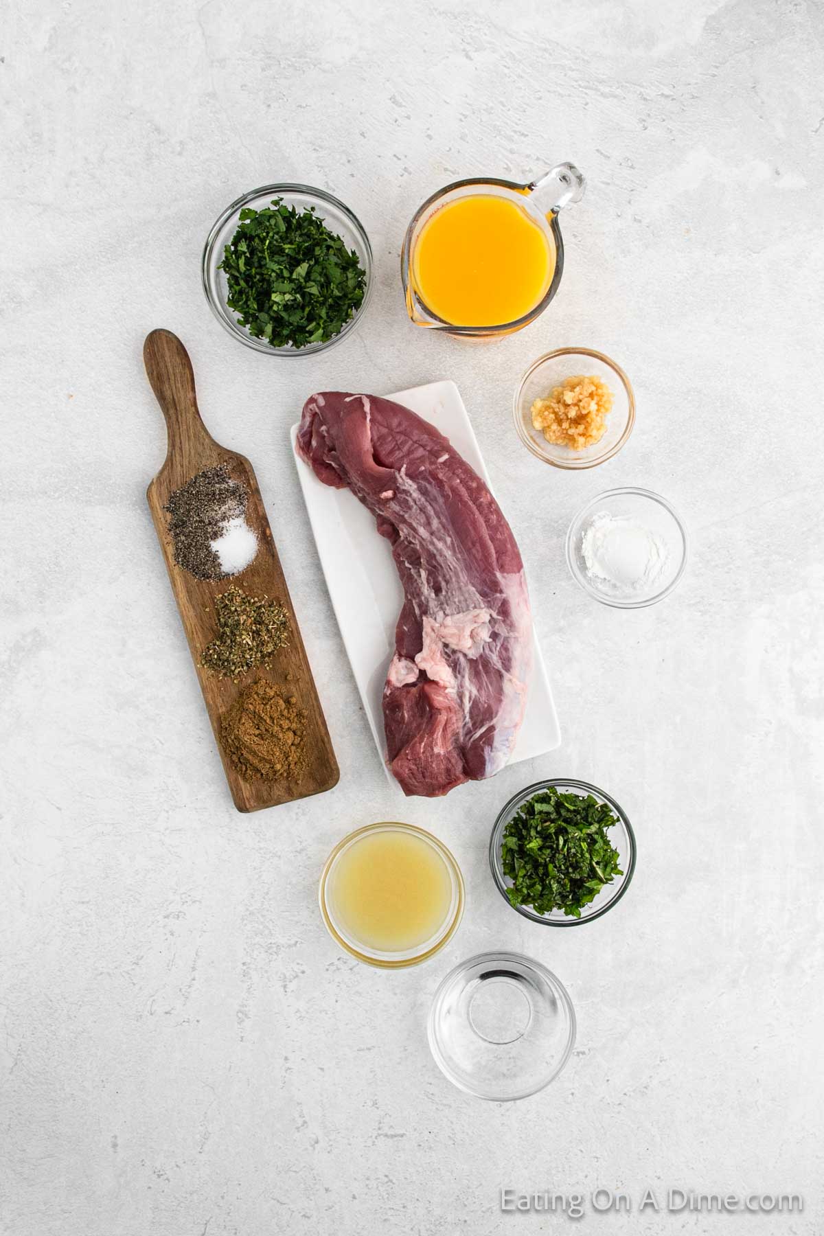 Top view of ingredients for a Crock Pot Cuban Mojo Pork Tenderloin recipe. A raw pork tenderloin sits on a white dish, surrounded by small bowls of chopped herbs, minced garlic, orange juice, lemon juice, oil, and seasonings. There is a wooden board with pepper, salt, and dried herbs.