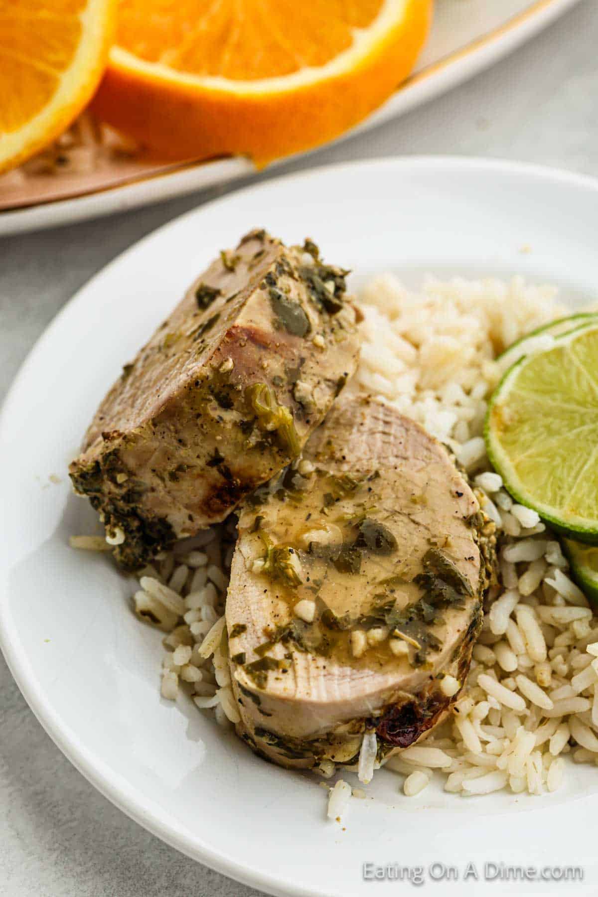 Two slices of seasoned pork tenderloin, inspired by a Crock Pot Cuban Mojo Pork Tenderloin recipe, are served on a bed of rice and garnished with lime slices on a white plate. In the background, orange slices rest on a separate dish. The pork features a green herb and seasoning coating.
