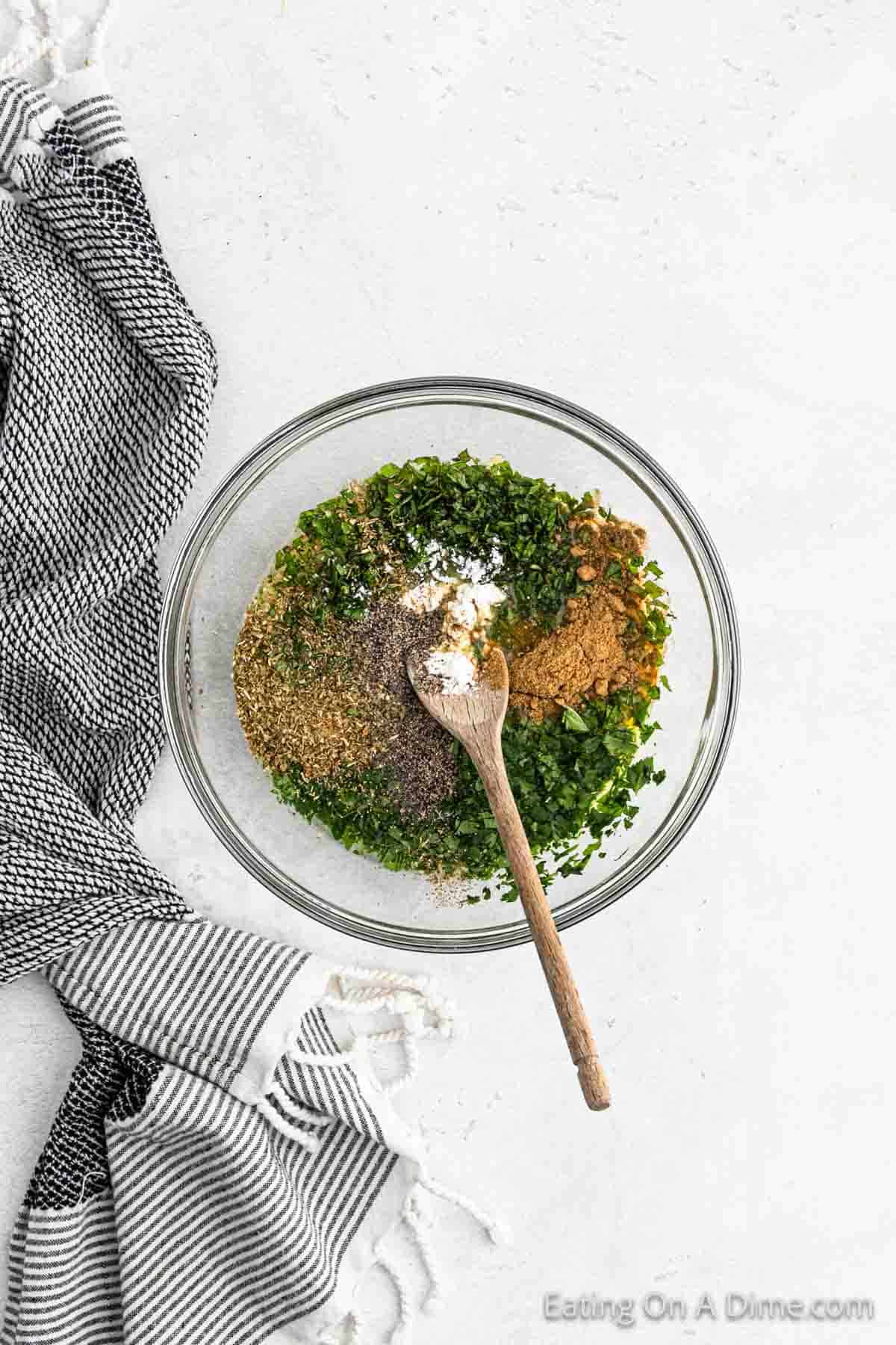 A clear glass bowl contains a blend of chopped herbs and spices, perfect for adding to a Crock Pot Cuban Mojo Pork Tenderloin recipe, with a wooden spoon resting inside. The bowl is placed on a light-colored surface next to a black and white striped kitchen towel.