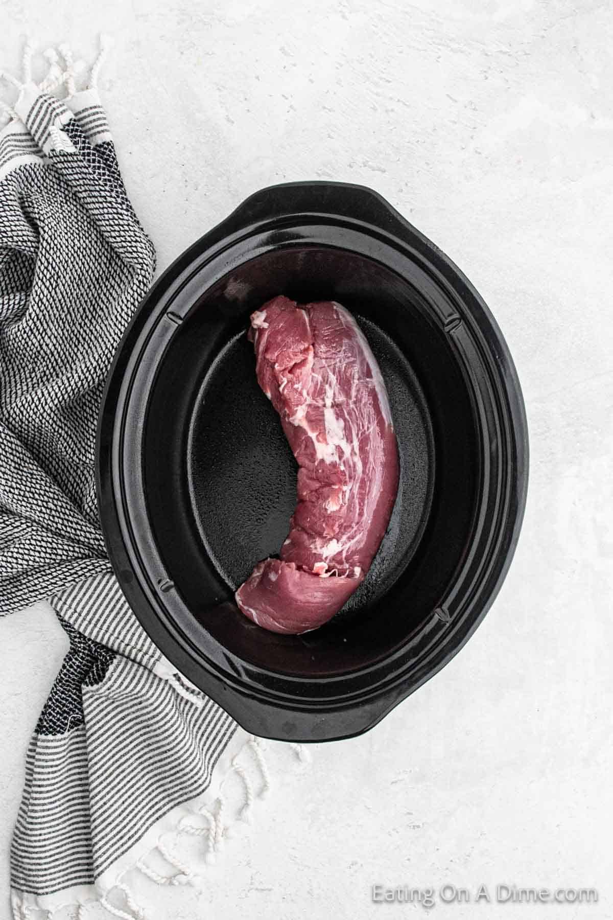 A raw pork tenderloin sits in a black slow cooker on a light gray surface, ready for the delicious Crock Pot Cuban Mojo Pork Tenderloin recipe. A white and dark gray striped kitchen towel is draped to the left of the slow cooker. The text "Eating On A Dime.com" is visible in the bottom right corner.