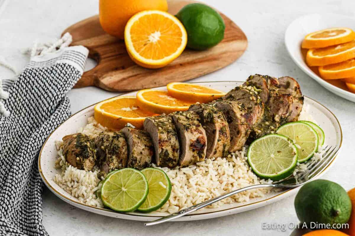 A platter of sliced, herb-crusted pork tenderloin rests on a bed of white rice, garnished with lime slices and accompanied by orange slices. Background includes a wooden board with a cut orange and whole lime, hinting at the flavors of our Crock Pot Cuban Mojo Pork Tenderloin recipe.