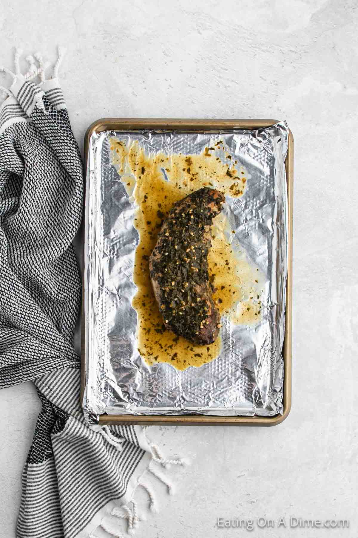 A seasoned and cooked steak rests on a foil-lined baking sheet, with juices pooling around it. The baking sheet is placed on a light gray surface next to a crumpled black and white striped cloth, reminiscent of the savory flavors found in a Crock Pot Cuban Mojo Pork Tenderloin recipe.
