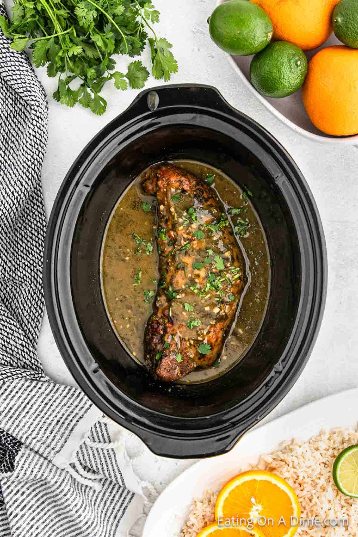 A cooked piece of meat with herbs and sauce is inside a black Crock Pot. Surrounding the slow cooker are a striped towel, a bunch of cilantro, a bowl of oranges and limes, and a plate of rice garnished with an orange slice, making it the perfect setting for a Cuban Mojo Pork Tenderloin recipe.