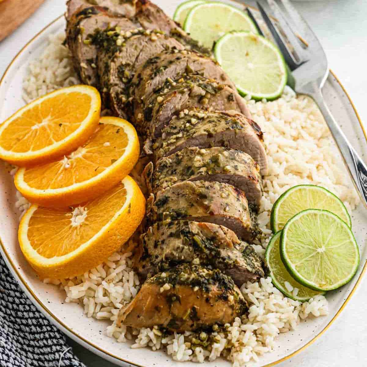 A plate of sliced seasoned Crock Pot Cuban Mojo Pork Tenderloin garnished with orange and lime slices, served over a bed of white rice. A serving fork is placed next to the tenderloin. The dish is presented on a white oval platter with a striped napkin nearby.