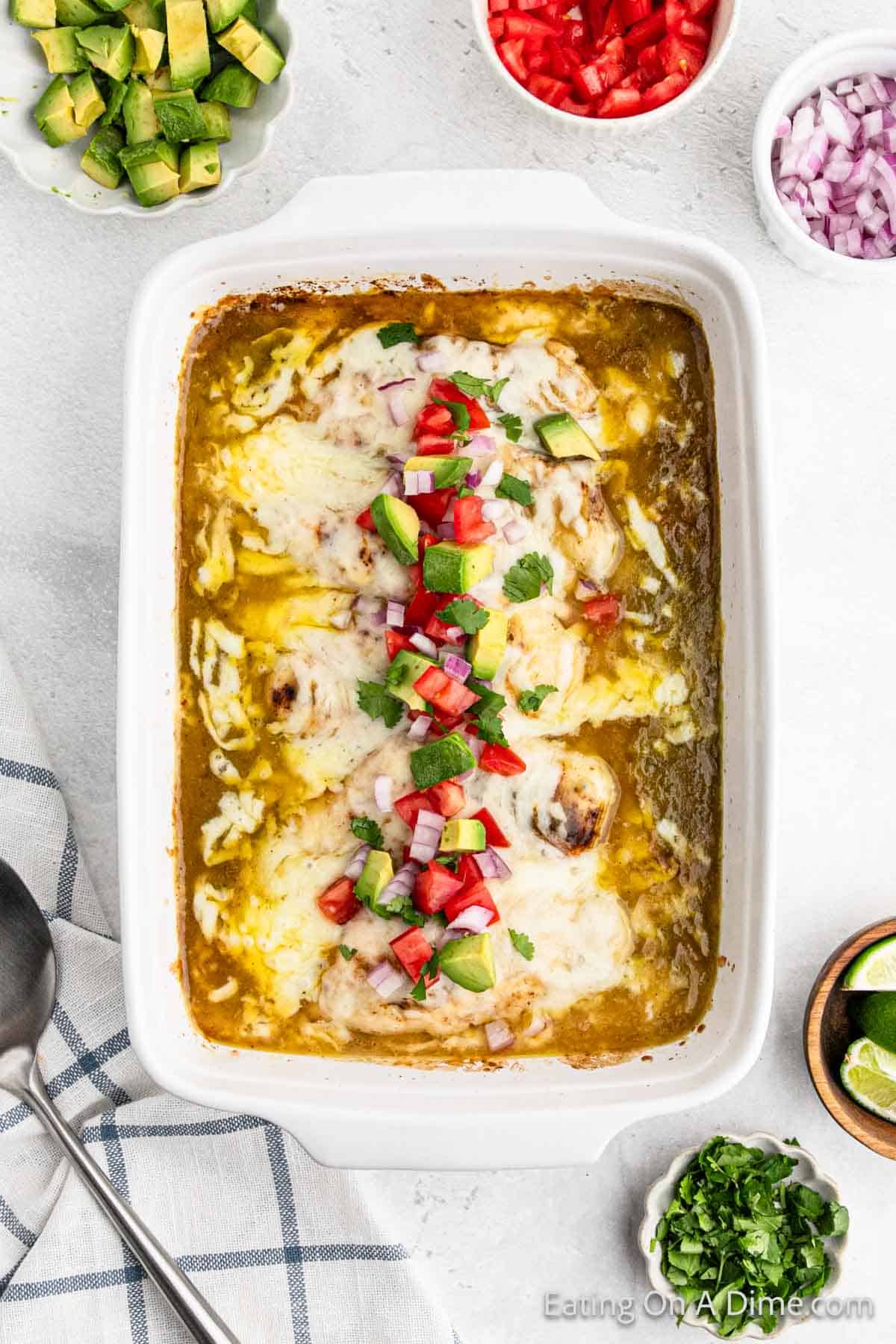 Baked enchilada chicken in a baking dish with enchilada sauce topped with melted cheese, diced avocado, tomato and fresh cilantro