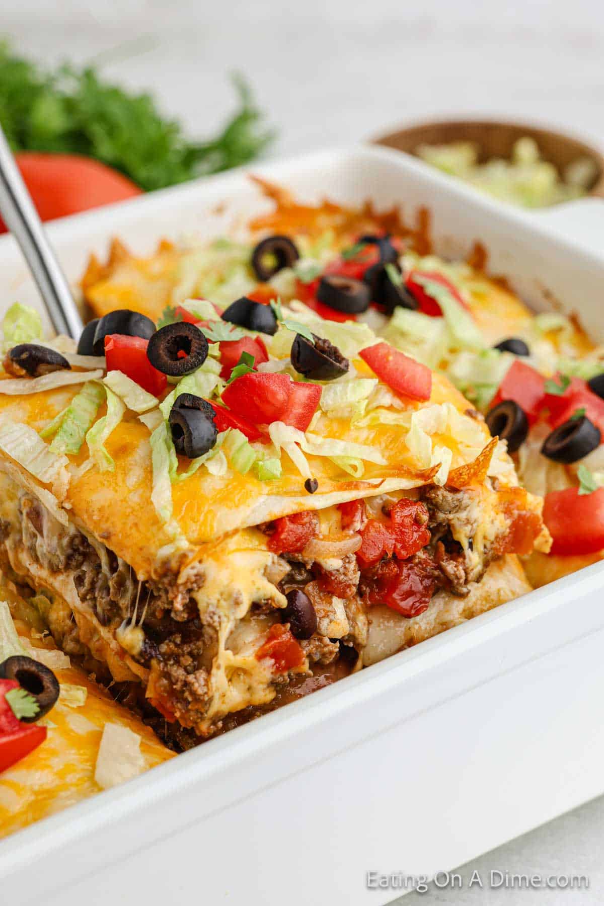 Taco Lasagna in a white casserole dish topped with lettuce, diced tomatoes and slice black olives