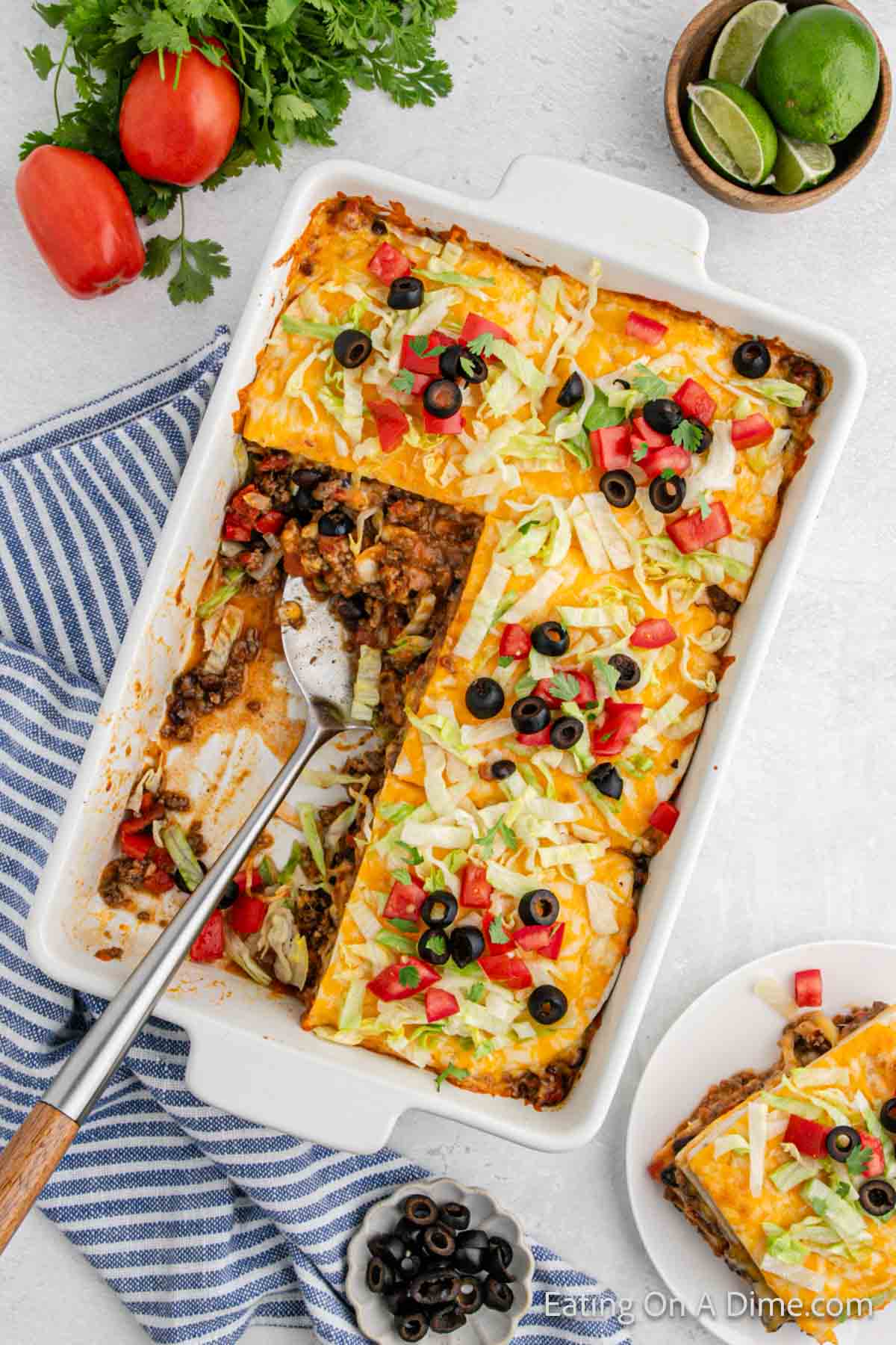 taco lasagna in a baking dish topped with shredded lettuce, slice black olives and diced tomatoes with empty space with a silver spatula