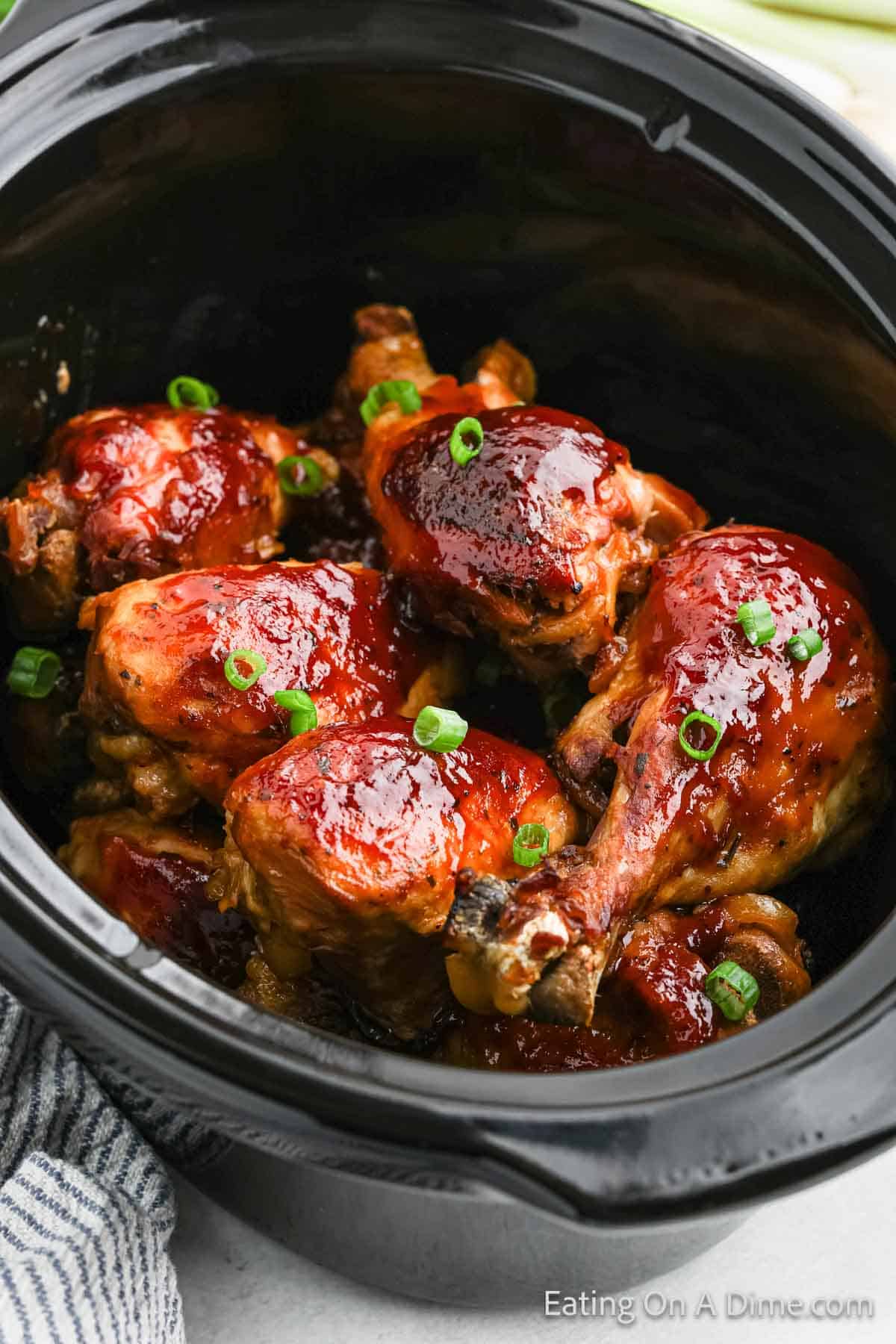 A crock pot filled with several cooked chicken drumsticks glazed with a rich, thick, red barbecue sauce, garnished with chopped green onions. The black interior of the crock pot contrasts with the vibrant colors of the dish. This BBQ ranch drumsticks recipe is perfect for a hearty family meal.