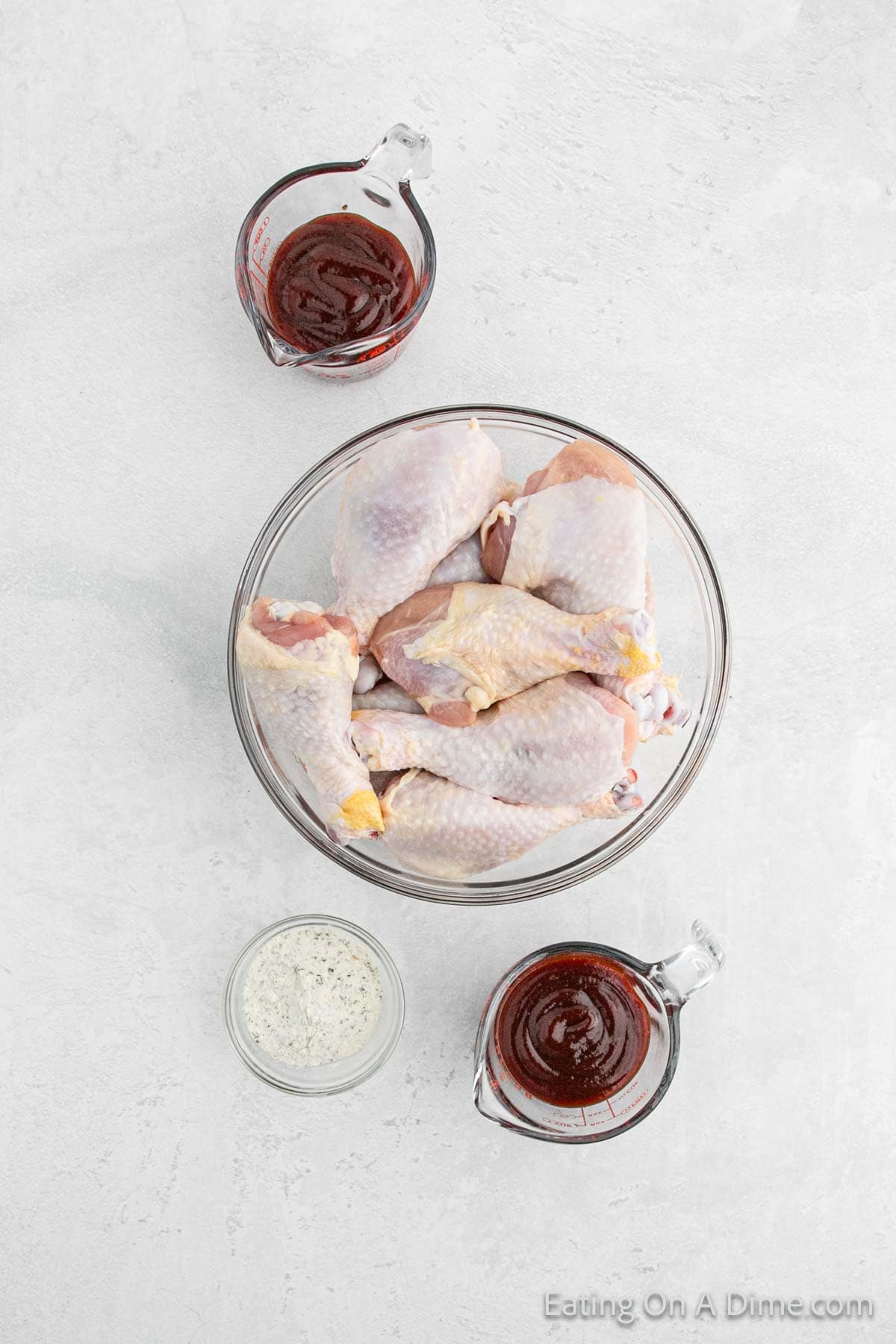 A glass bowl filled with raw chicken pieces is at the center. Surrounding the bowl are three glass containers: one with BBQ sauce on top, another with a dry white seasoning mix at the bottom, and a third with more BBQ sauce on the right, setting the stage for a delicious Crock Pot BBQ Ranch Drumsticks recipe.