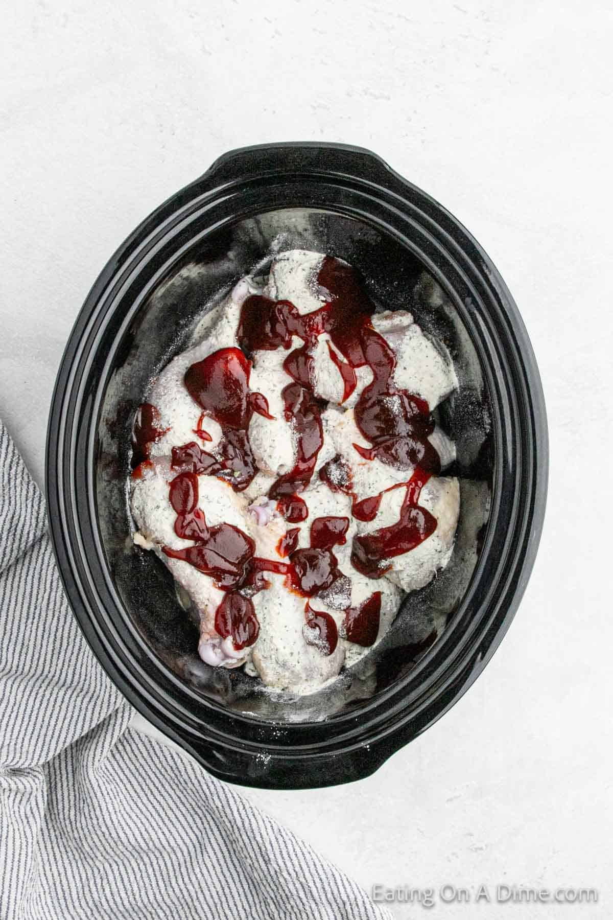 A black crock pot filled with chicken breasts covered in a creamy white sauce and drizzled with a reddish-purple sauce. The cooker is set on a light-colored countertop next to a folded, white and gray striped cloth, reminiscent of a delicious BBQ ranch recipe but tailored for tender chicken.