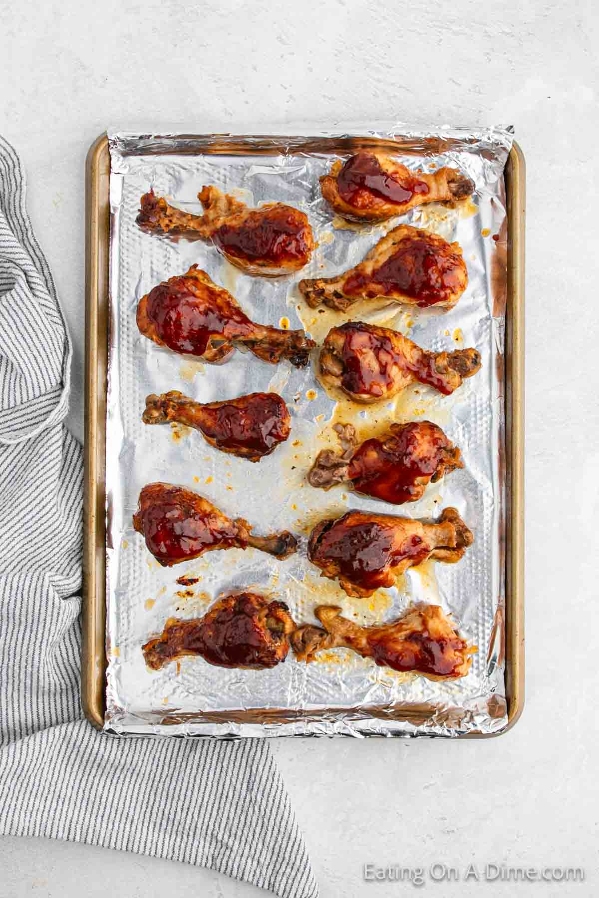 A baking sheet lined with aluminum foil is filled with 12 Crock Pot BBQ Ranch drumsticks covered in barbecue sauce. The sheet is placed on a light-colored surface next to a striped cloth. The text "Eating On A Dime.com" is visible in the bottom right corner.