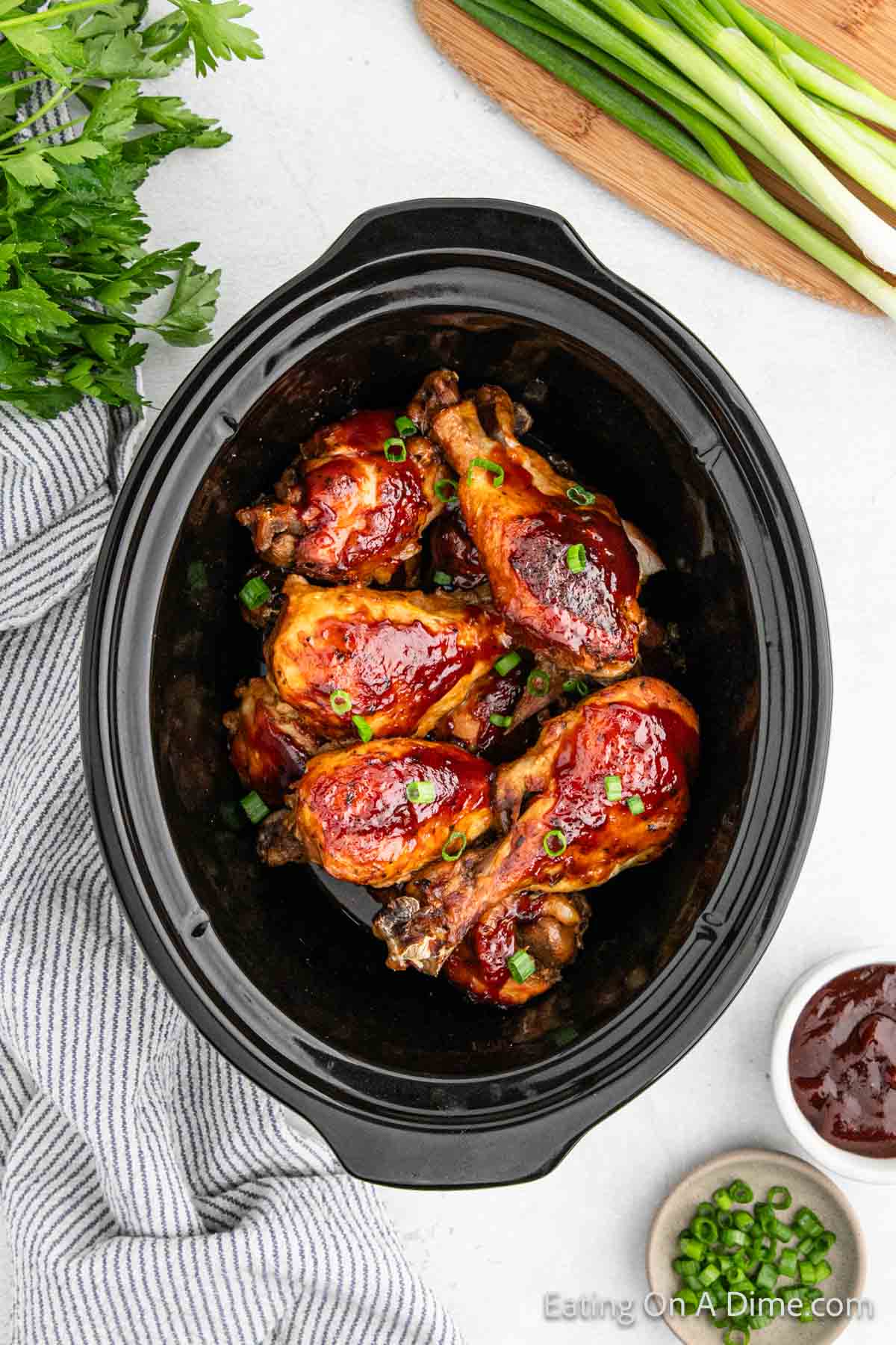 A close-up view of a crock pot filled with BBQ ranch drumsticks, garnished with chopped green onions. The slow cooker is on a white surface, surrounded by fresh parsley, green onions, and a striped kitchen towel. A small bowl of sauce is next to the cooker.