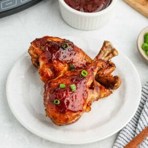 Two juicy, glazed crock pot BBQ ranch drumsticks garnished with chopped green onions are served on a white plate. A small bowl of barbecue sauce and a dish with additional chopped green onions are in the background. The setting includes a striped cloth napkin to the side.