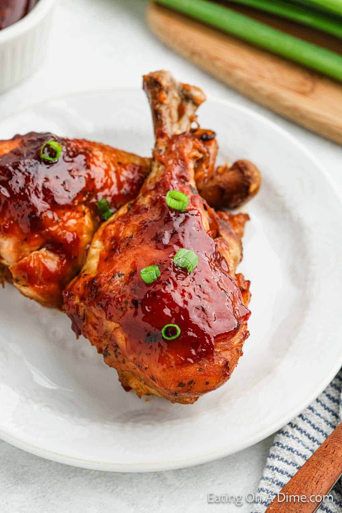 A close-up of a plate with two glazed chicken legs garnished with sliced green onions. The chicken legs appear juicy with a shiny, caramelized sauce, reminiscent of a crock pot BBQ ranch drumsticks recipe. A striped cloth napkin is partially visible in the corner.