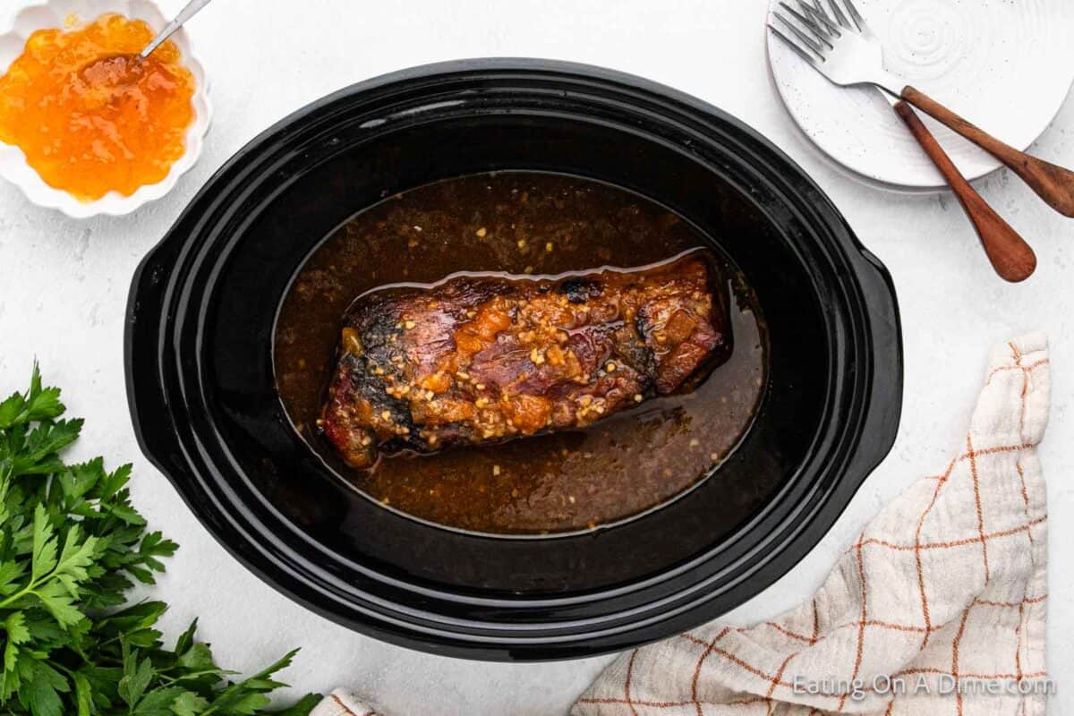 A beef roast glistens with glaze in a black slow cooker, reminiscent of a Crock Pot Pork Tenderloin with Peaches. Beside it, an orange sauce awaits with a spoon, forks rest on a white saucer, and a green herb garnish adds freshness. A light cloth napkin is casually draped at the bottom right.