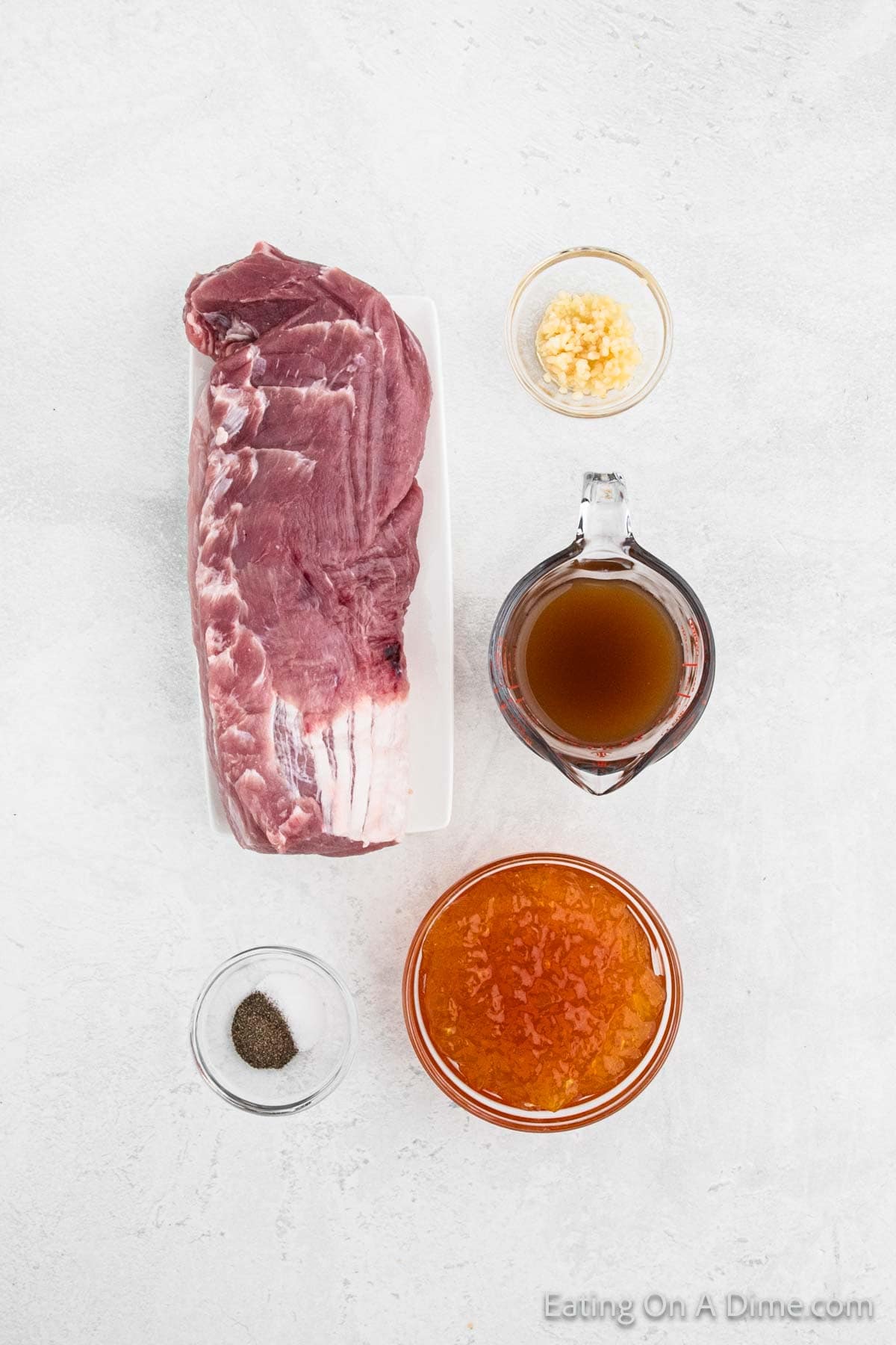 Top view of a raw pork tenderloin on a white plate, surrounded by small bowls of chopped garlic, brown sauce, orange marmalade, and a dish of black pepper on a light gray background—perfect for creating Crock Pot Pork Tenderloin with Peaches.