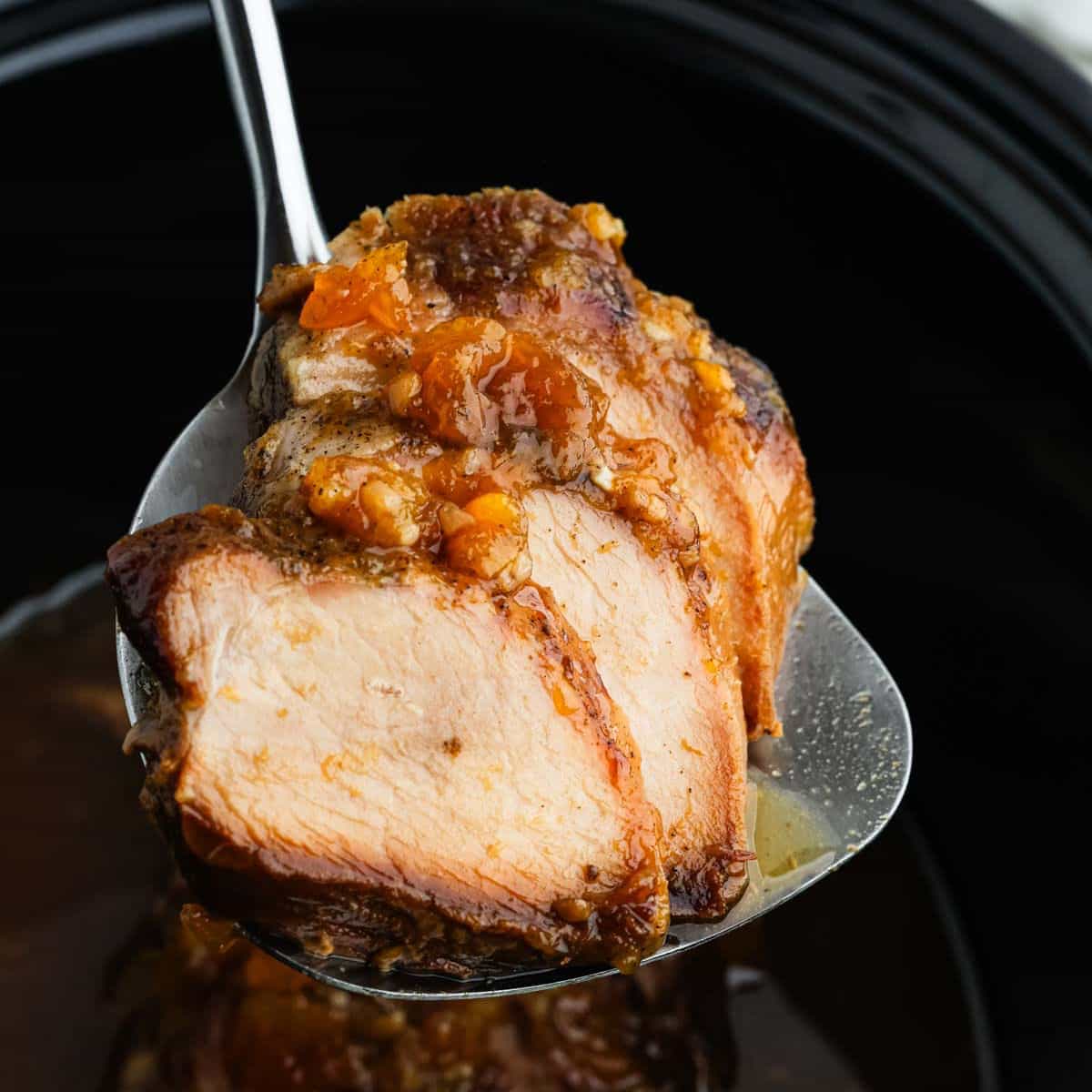 A slice of tender Crock Pot Pork Tenderloin with a sweet glaze and garnished with small bits of herbs and spices, accompanied by juicy peaches, held on a spoon. The background is slightly blurred, emphasizing the succulent texture and rich color of the meat.