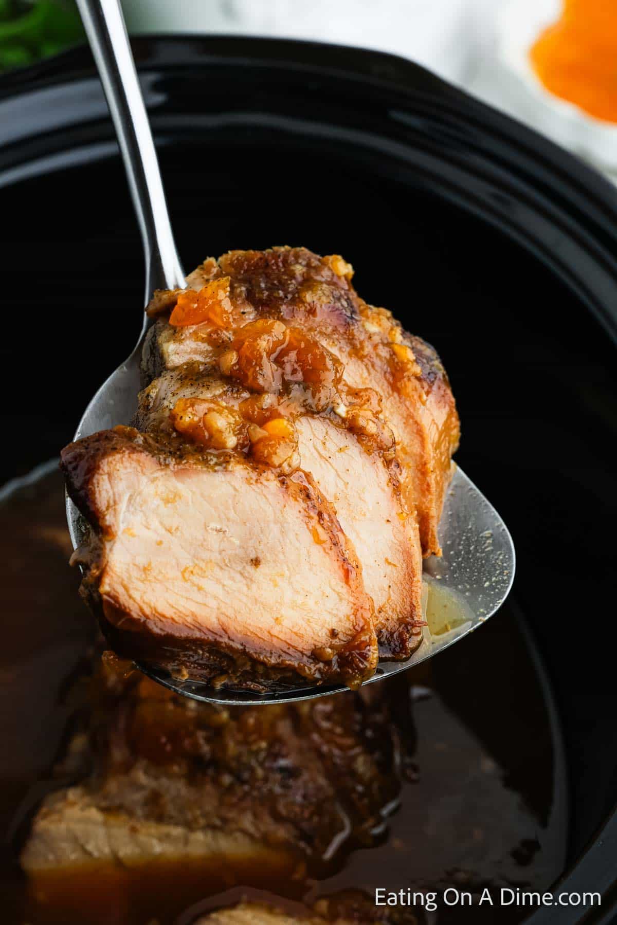 A close-up of a spoon holding a slice of pot roast with visible spices and juices, similar to how crock pot pork tenderloin with peaches might look. The slice is poised over a slow cooker containing more savory pot roast. In the background, an orange and white item peeks through.