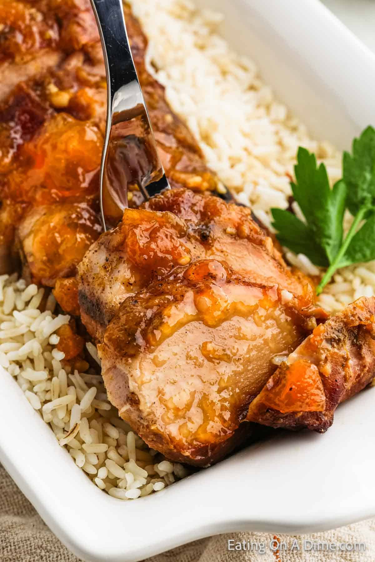 A fork slices into glazed, roasted pork tenderloin, reminiscent of Crock Pot Pork Tenderloin with Peaches, placed on a bed of white rice in a rectangular dish. A sprig of fresh parsley decorates the dish, adding a pop of green next to the golden-brown meat.