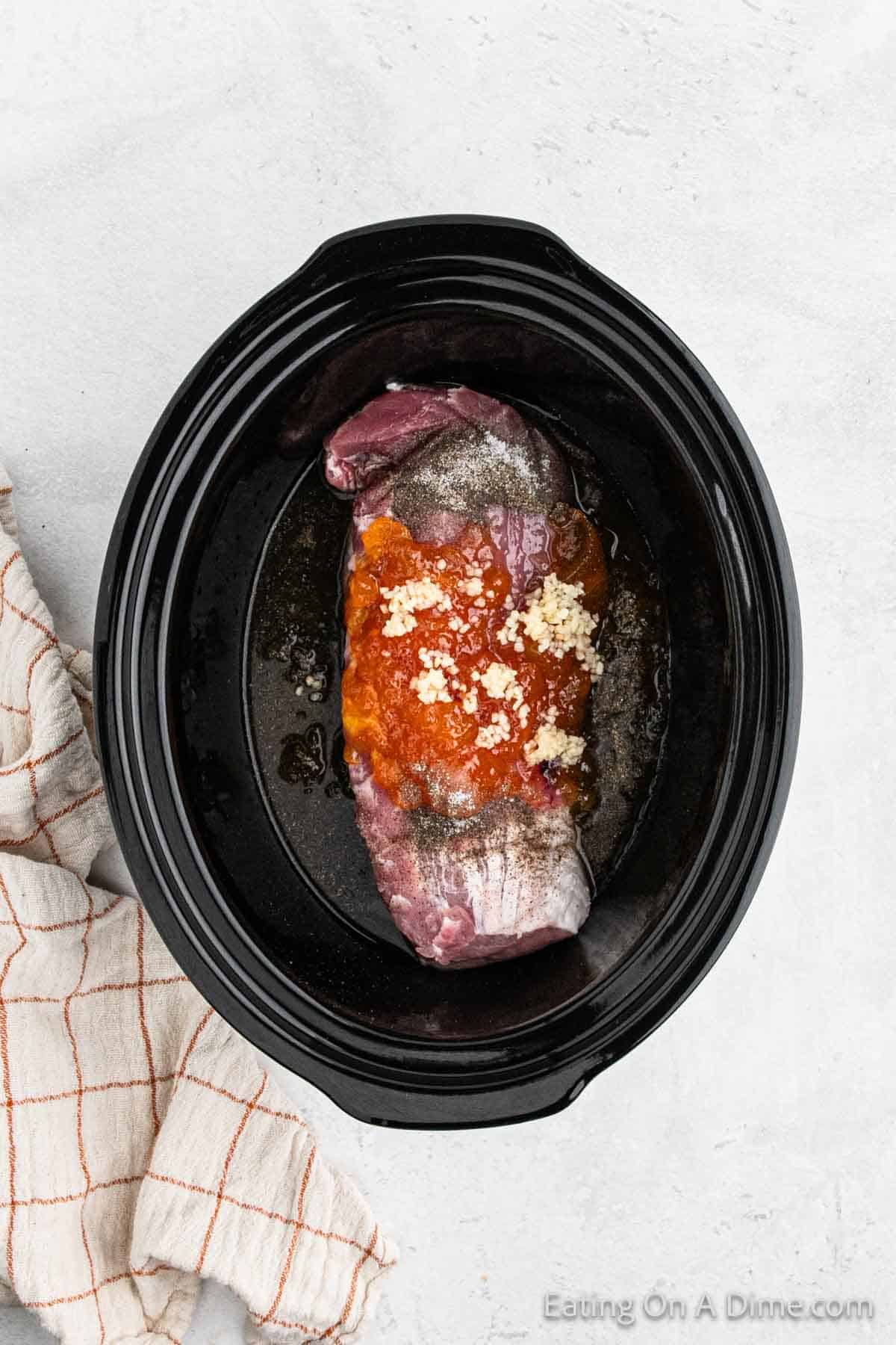 A Crock Pot Pork Tenderloin with peaches sits marinating in sauce and minced garlic. The slow cooker rests on a light gray surface, accompanied by a white and orange checkered cloth, promising a savory blend of flavors.